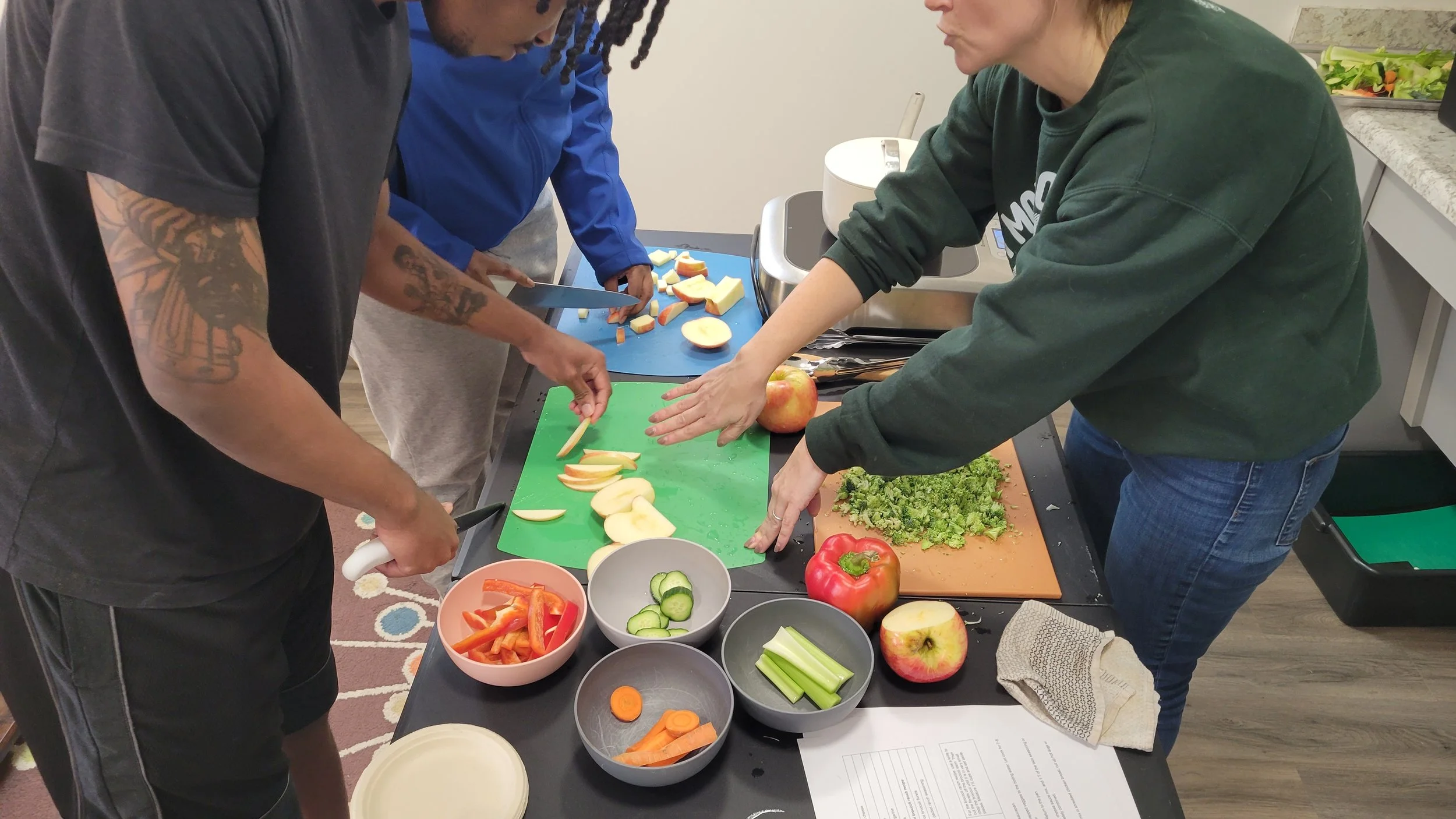 Kait of Burkett Farm gestures while two young adults cut apples into slices. Also on the table is a cutting board full of chopped broccoli and bowls of cut raw veggies including bell peppers, cucumbers, celery, and carrots.