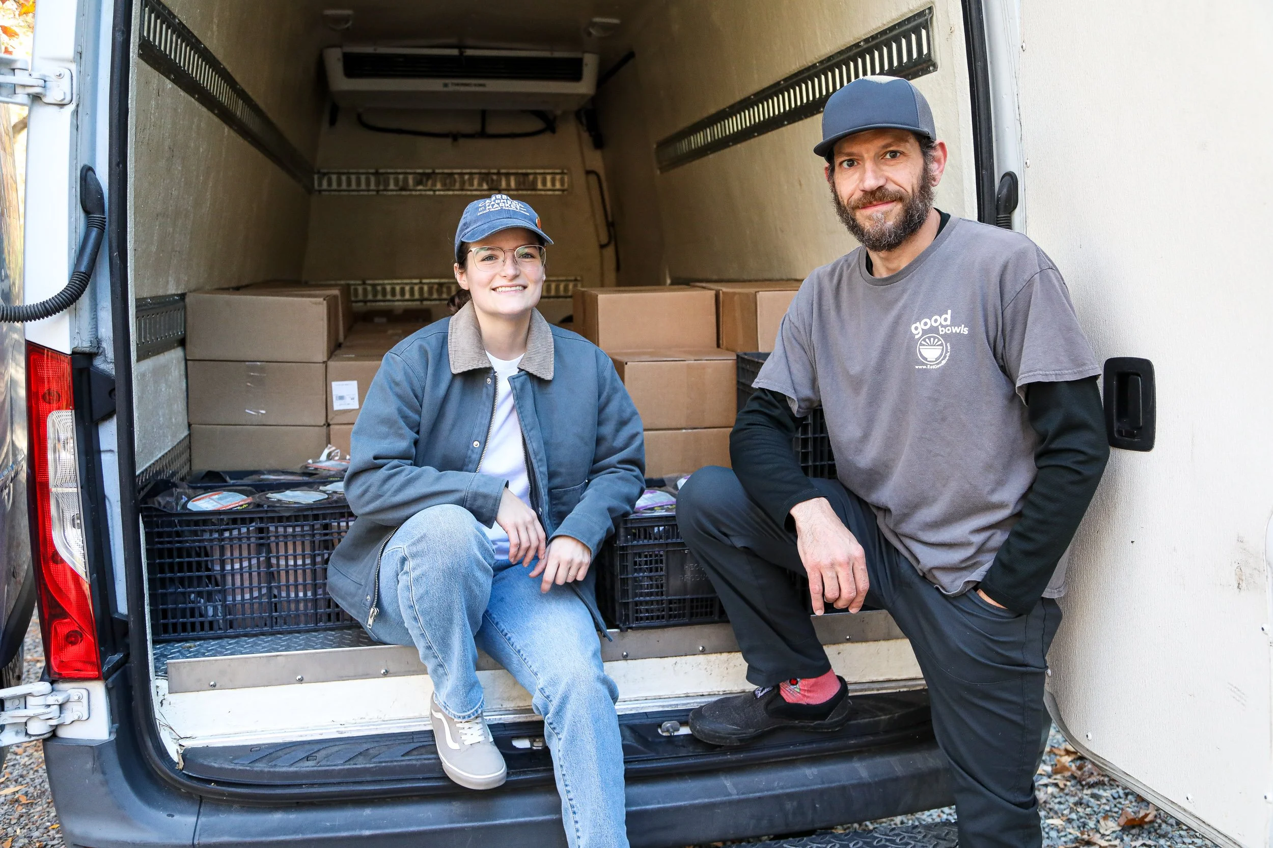 A woman, Tori Wierzchowski, Executive Director of Food Equity Collaborative, and a man, Steve Navarra of Equiti Foods, sit in the back of an open van. Many cardboard boxes and plastic crates of Good Bowls sit in the van behind them.