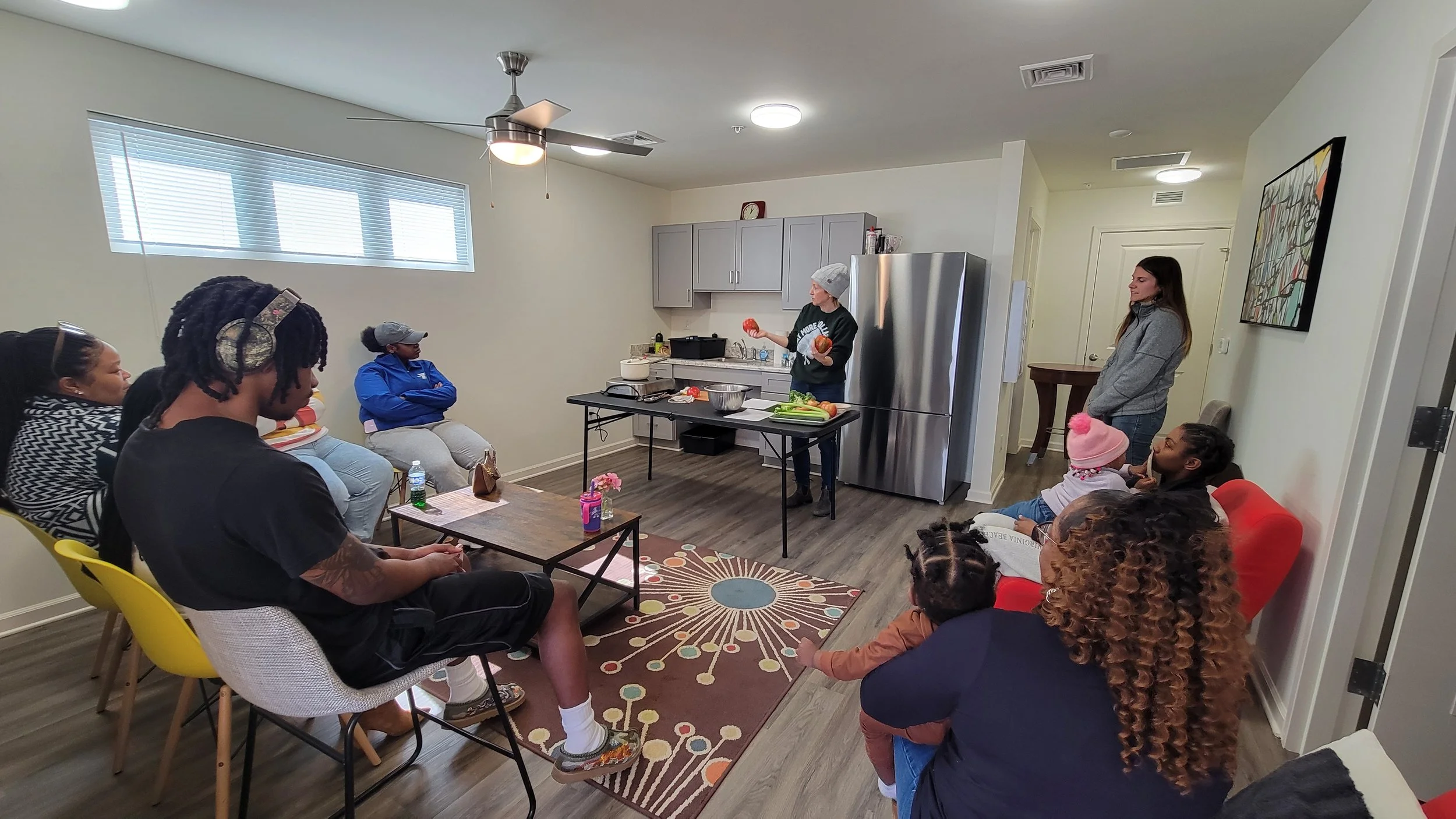 View of a small room with multiple people seated looking at a table in the center of the room. Kait of Burkett Farm stands at the table holding vegetables while doing a cooking demonstration.