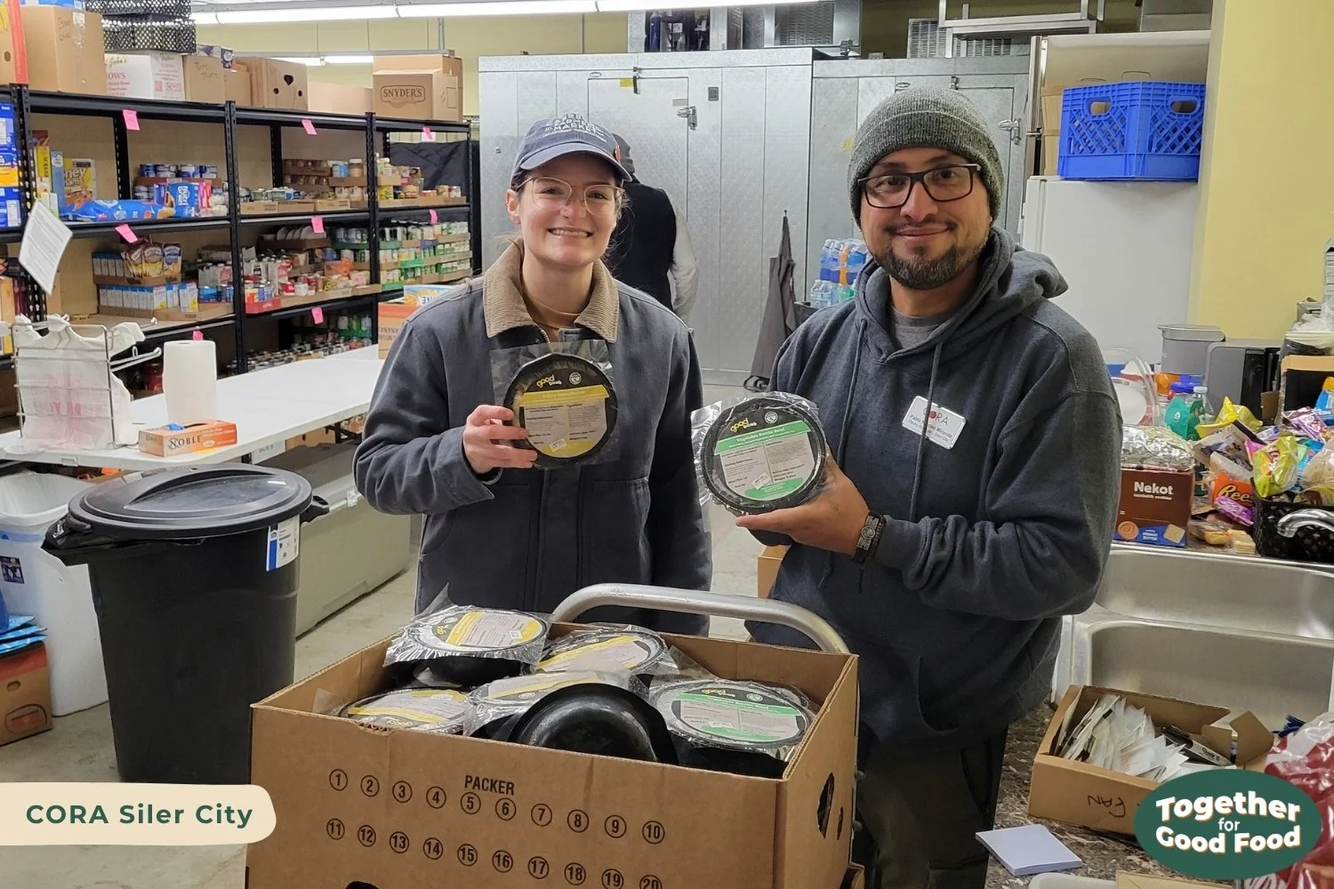 Tori Wierzchowski of Equiti Foods and Pablo of CORA Siler City stand in the food pantry behind a cart of Good Bowls. Both are smiling and holding up a Good Bowl. 