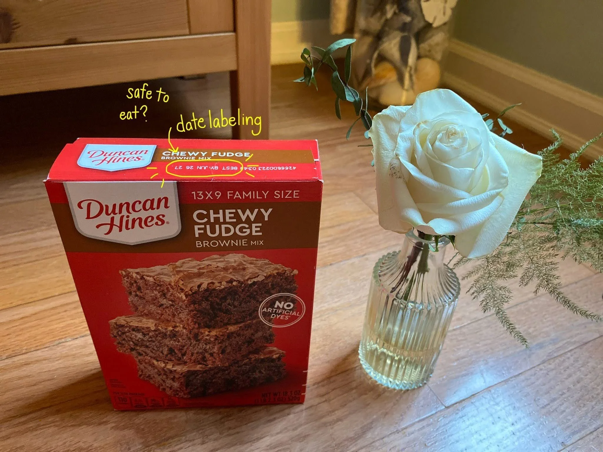 A brownie mix box next to one white rose in a vase. The food date label is circled in yellow with “date labeling” and “safe to eat?” handwritten in yellow text.