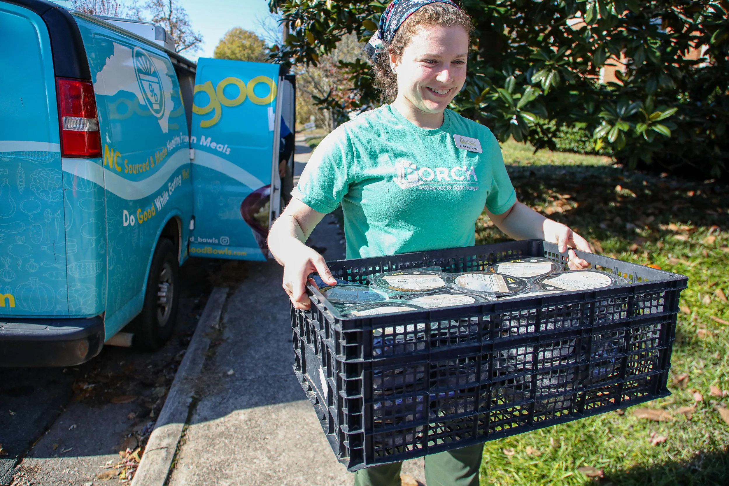 In the foreground, a PORCH staff member is smiling and walking with a crate full of Good Bowls. The Good Bowls delivery van is in the background.