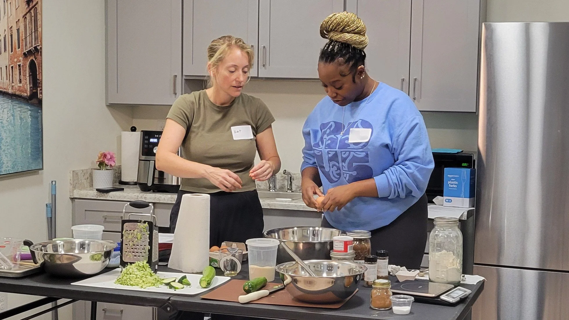 Kait of Burkett Farm and a Kitchen Compass client crack eggs into a mixing bowl at a table full of zucchini muffin ingredients. 