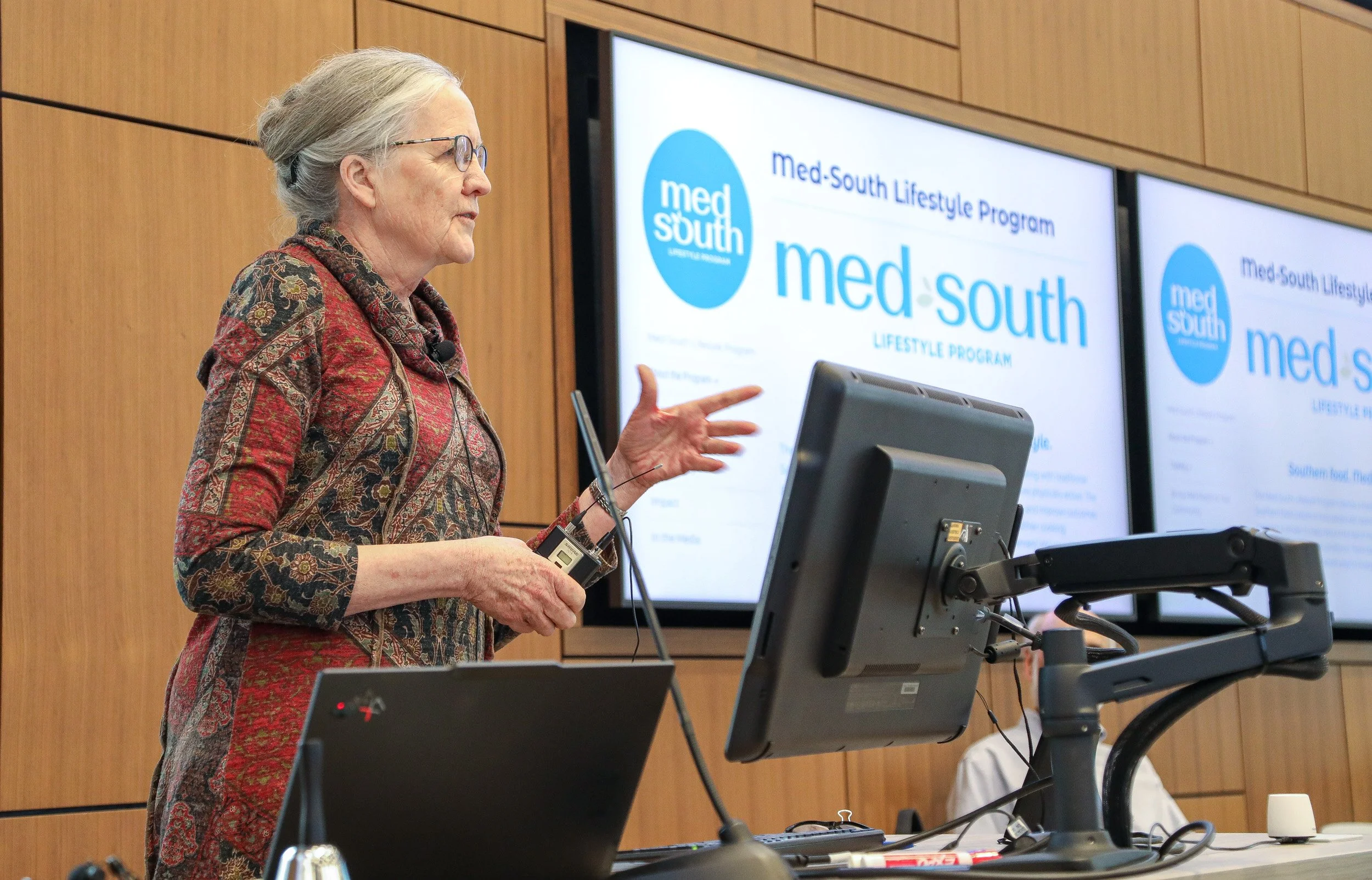 Alice Ammerman standing behind a desk with a computer. She is gesturing while presenting to a crowd beyond the image. In the background is a slideshow on a large TV projecting information about Med-South.