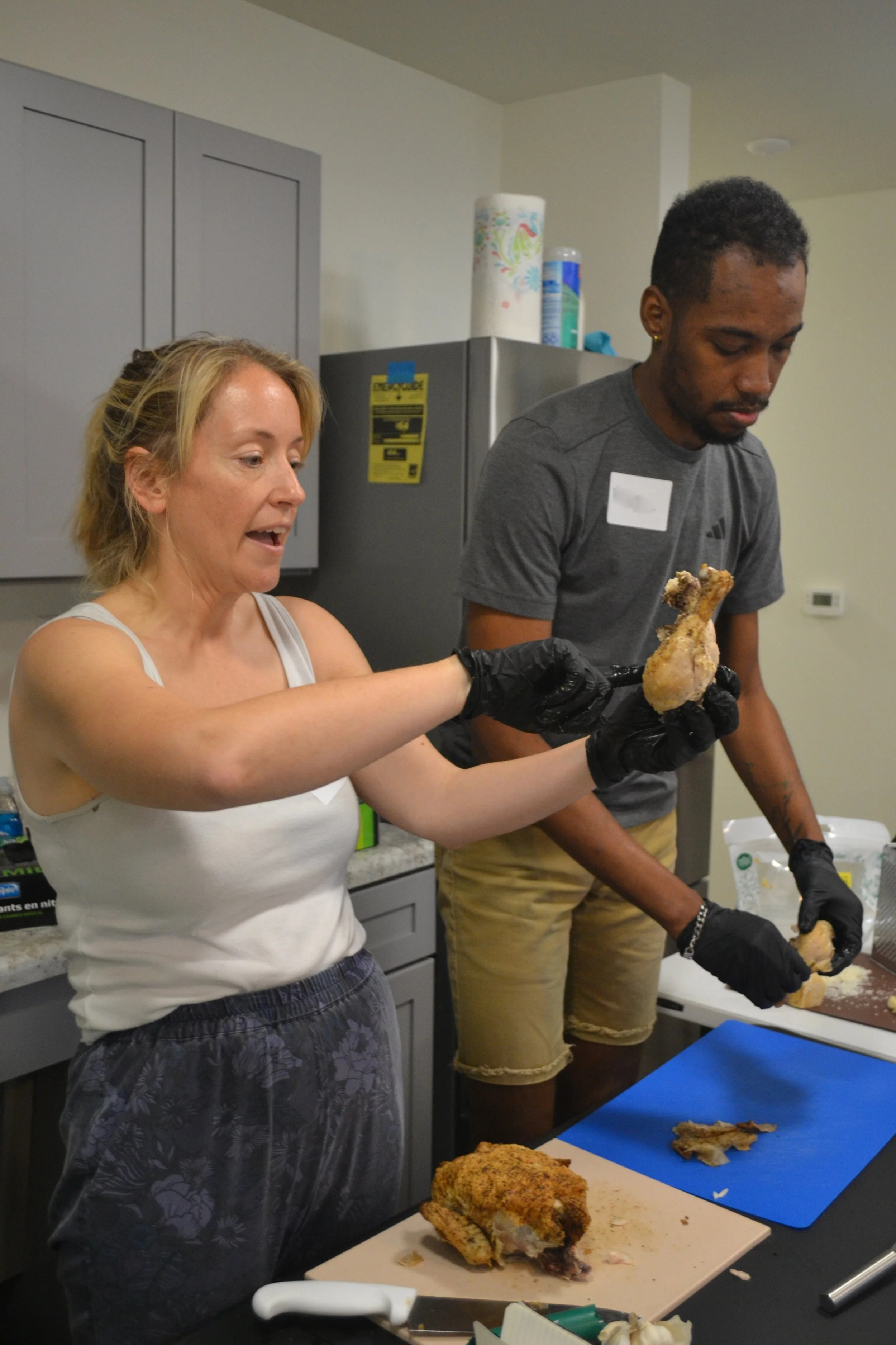Kait of Burkett Farm holding a piece of rotisserie chicken while explaining how to remove meat from the bone. A Kitchen Compass client is removing meat from another piece in the background.