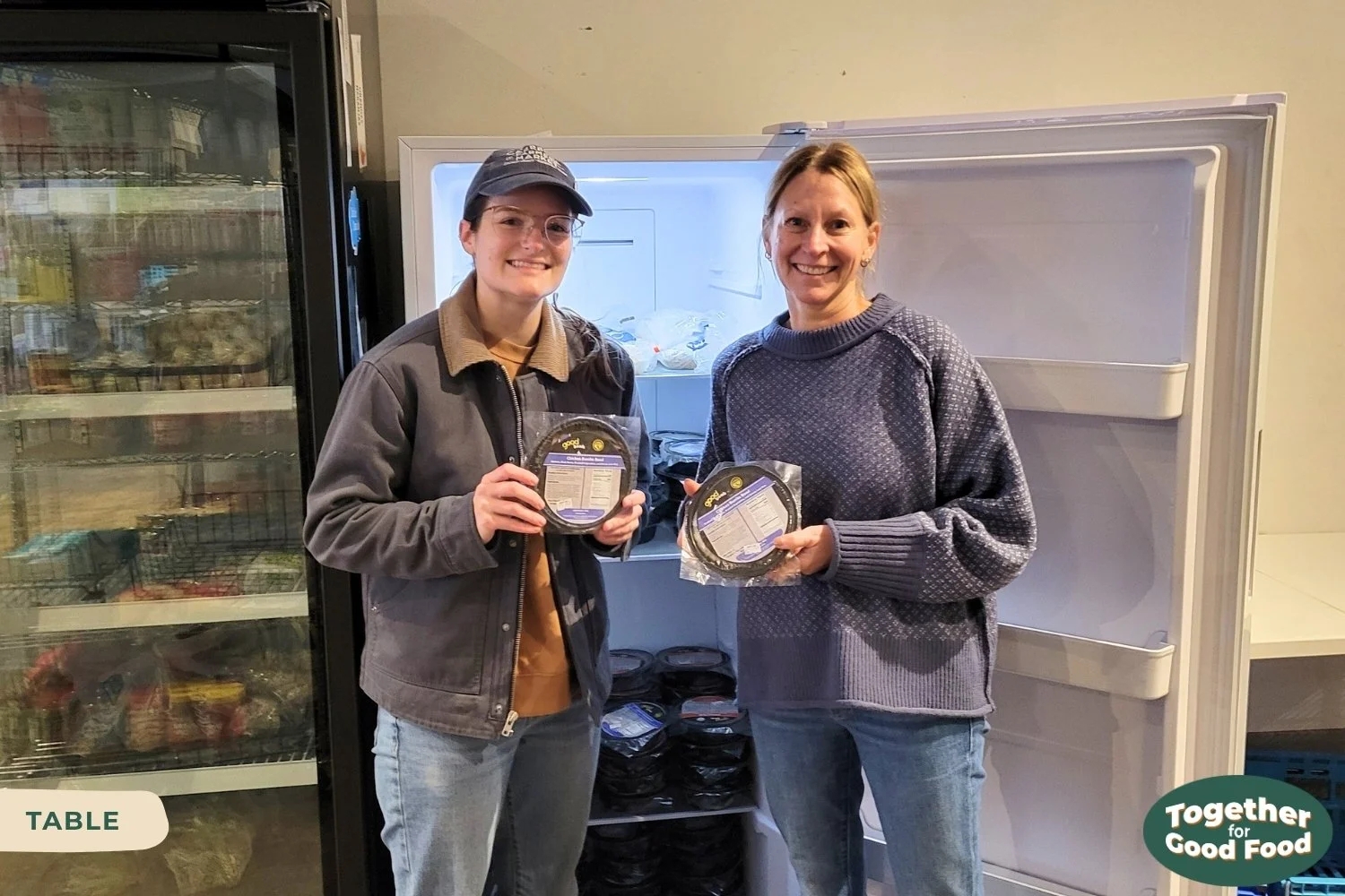 Tori Wierzchowski of Food Equity Collaborative and Suzanne of TABLE standing in front of an open freezer at TABLE, each holding a Good Bowl and smiling.