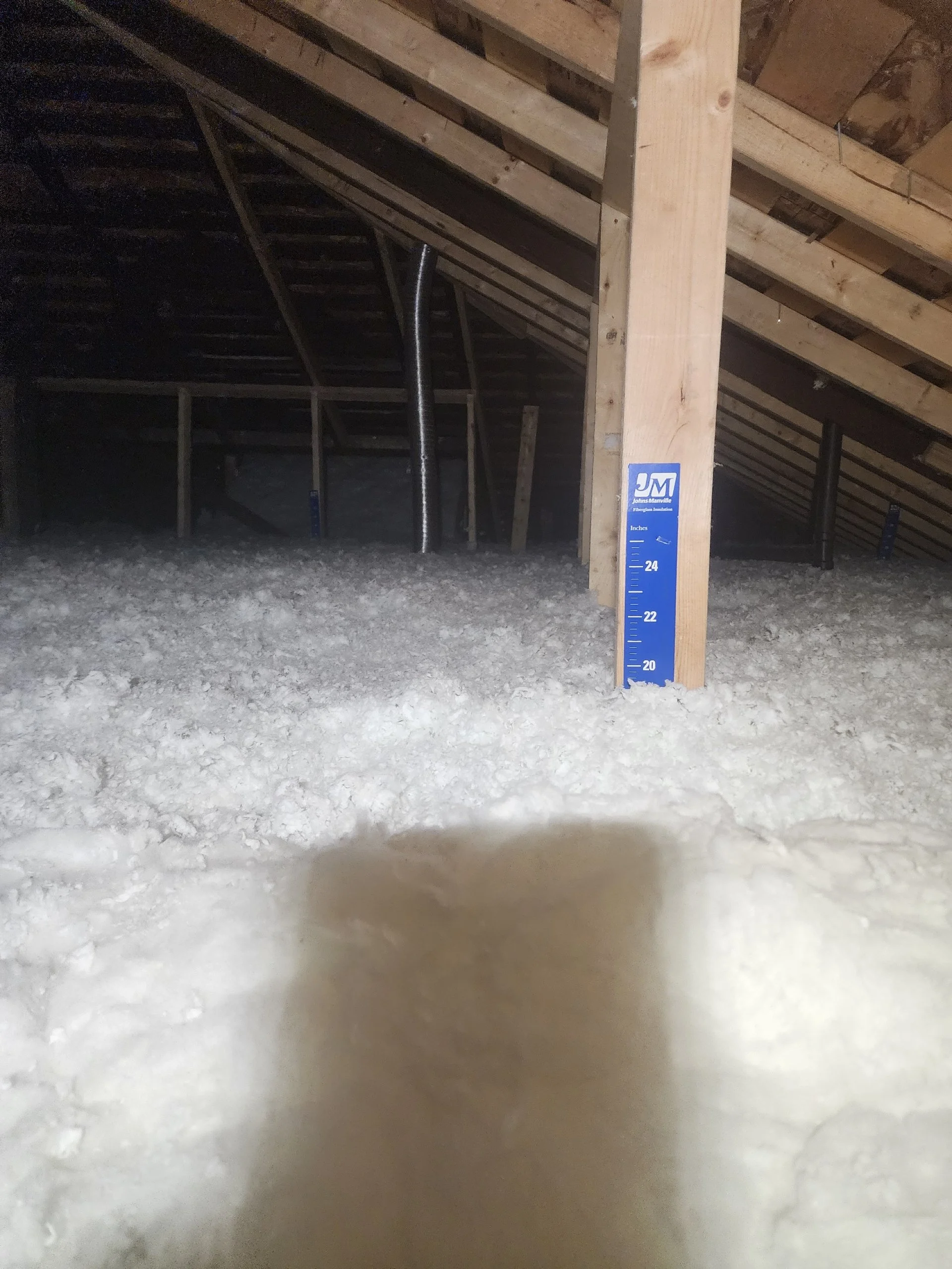 View of an attic with exposed wooden beams, insulation, and a blue measuring stick indicating height, with some pipes and a sloped ceiling.