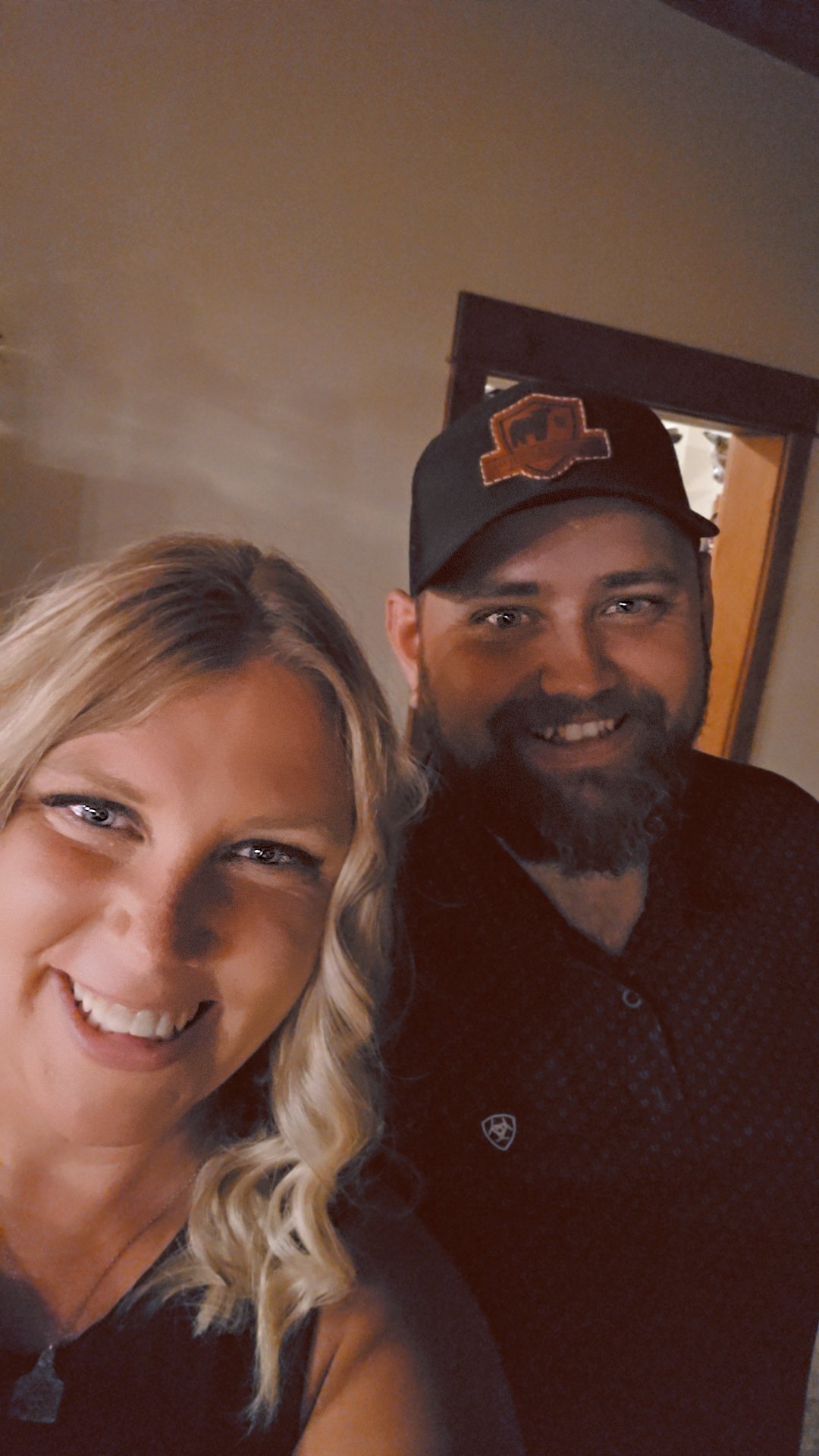 A smiling woman with blonde hair and a bearded man wearing a black cap. They are taking a selfie together indoors.