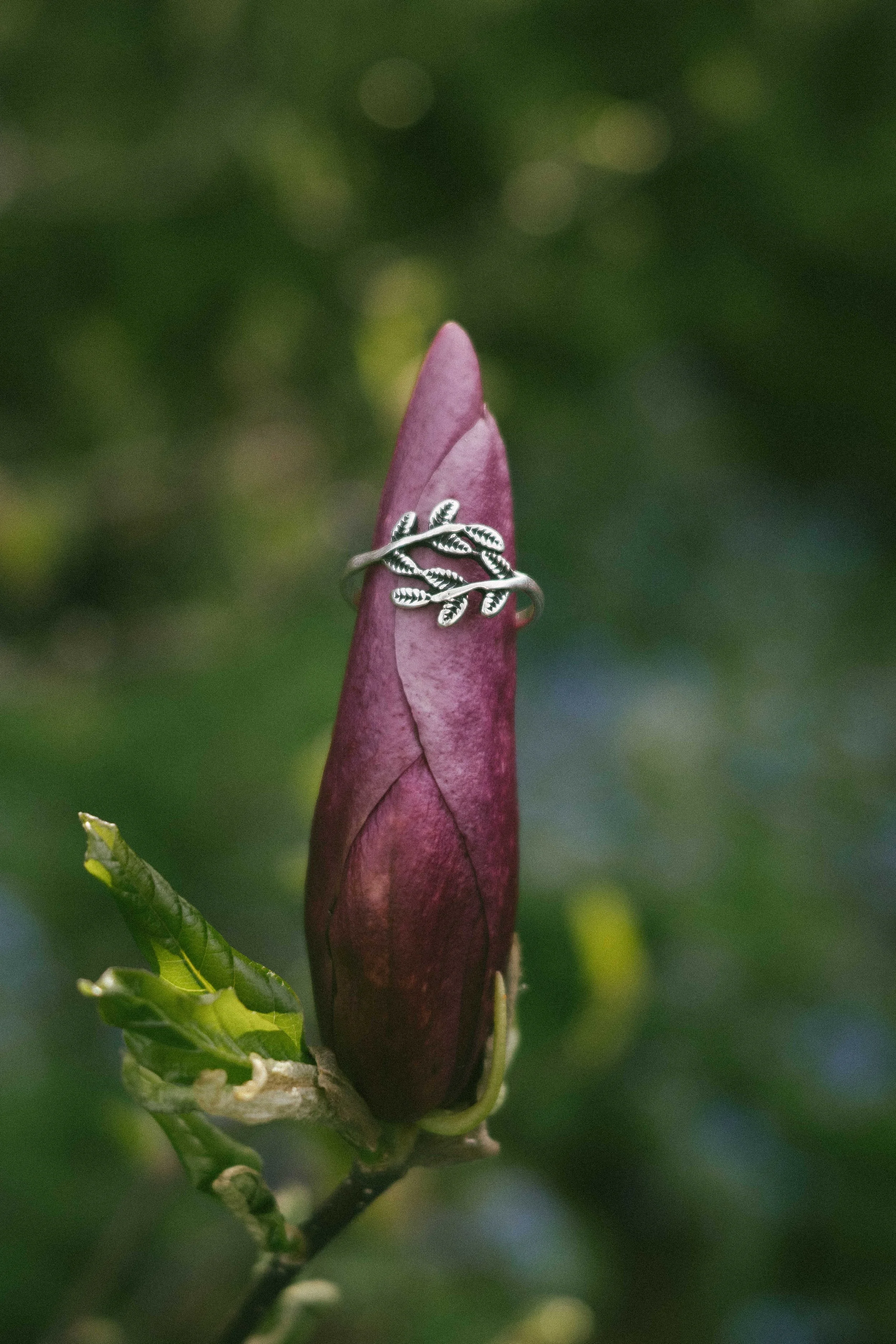 Close-up of a silver leaf-inspired ring wrapped around a deep purple flower bud, photographed outdoors with a soft green background. Nature-inspired botanical jewelry with an organic, handcrafted aesthetic, perfect for boho style and artisan silver c