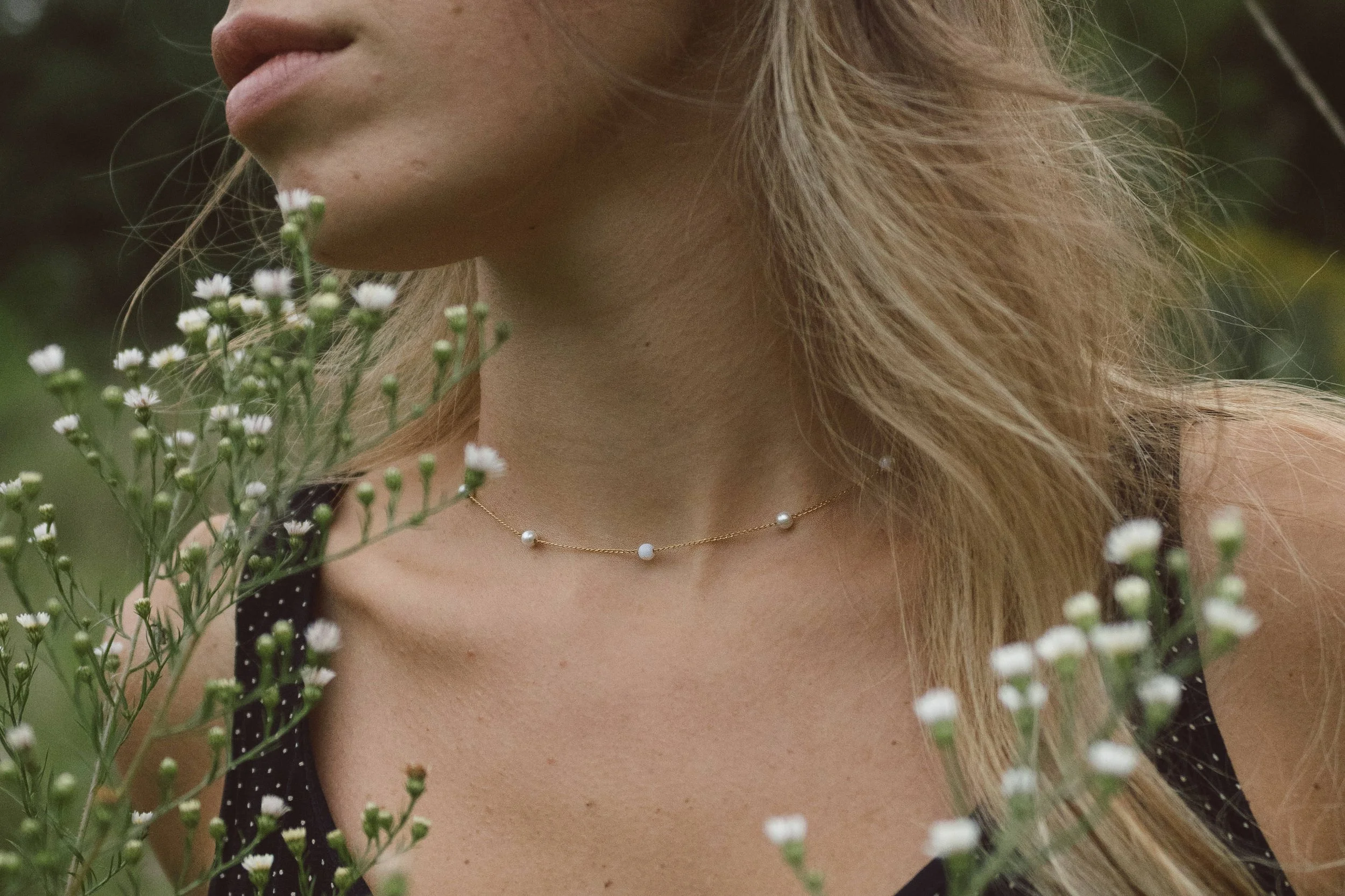 Lifestyle portrait of a woman wearing a minimalist gold pearl necklace, standing outdoors among wildflowers. Delicate white pearls rest along the collarbone, styled with a natural, romantic aesthetic—ideal for bridal jewelry, everyday elegance, and n
