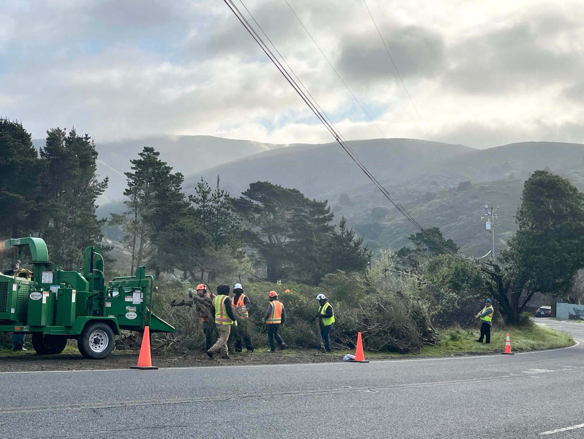 People working on a roadside repair or maintenance project with traffic cones set up, a green utility vehicle, and trees with hills and cloudy sky in the background.