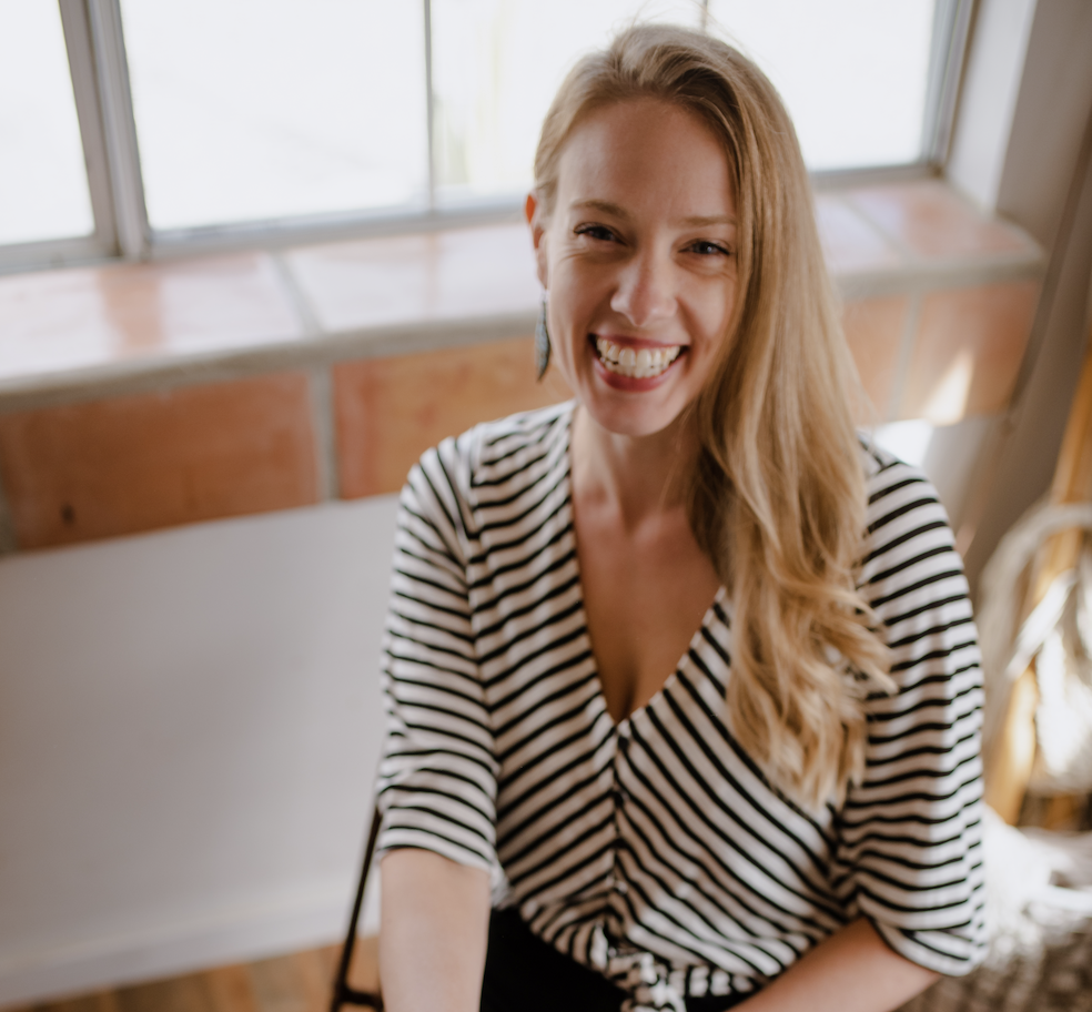 A woman with long, wavy blonde hair smiling at the camera, wearing a black and white striped top, indoors near a window.