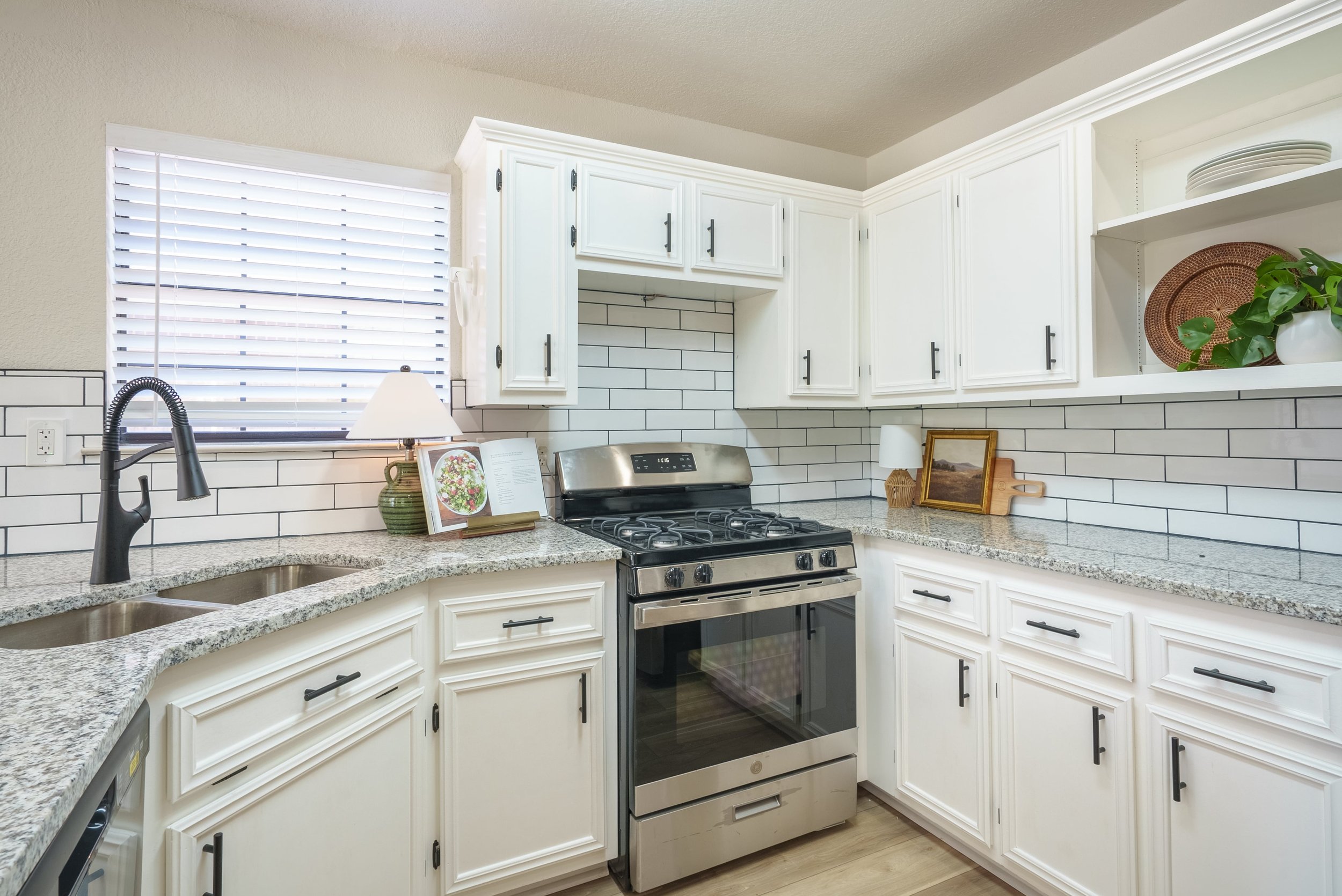 Modern kitchen with white cabinets, granite countertops, stainless steel stove, subway tile backsplash, double sink, and decorative elements like a plant and lamp.