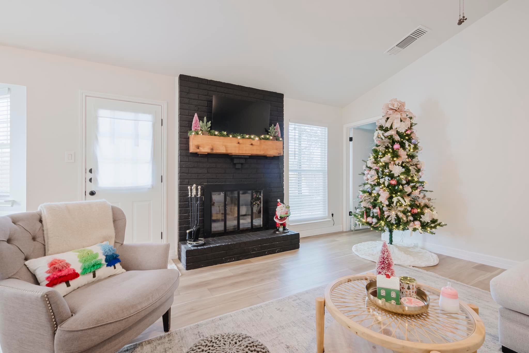 Living room with a Christmas theme, including a decorated Christmas tree, a black fireplace with a TV and festive garland, a cozy armchair with a colorful pillow, and a round coffee table with holiday decorations.