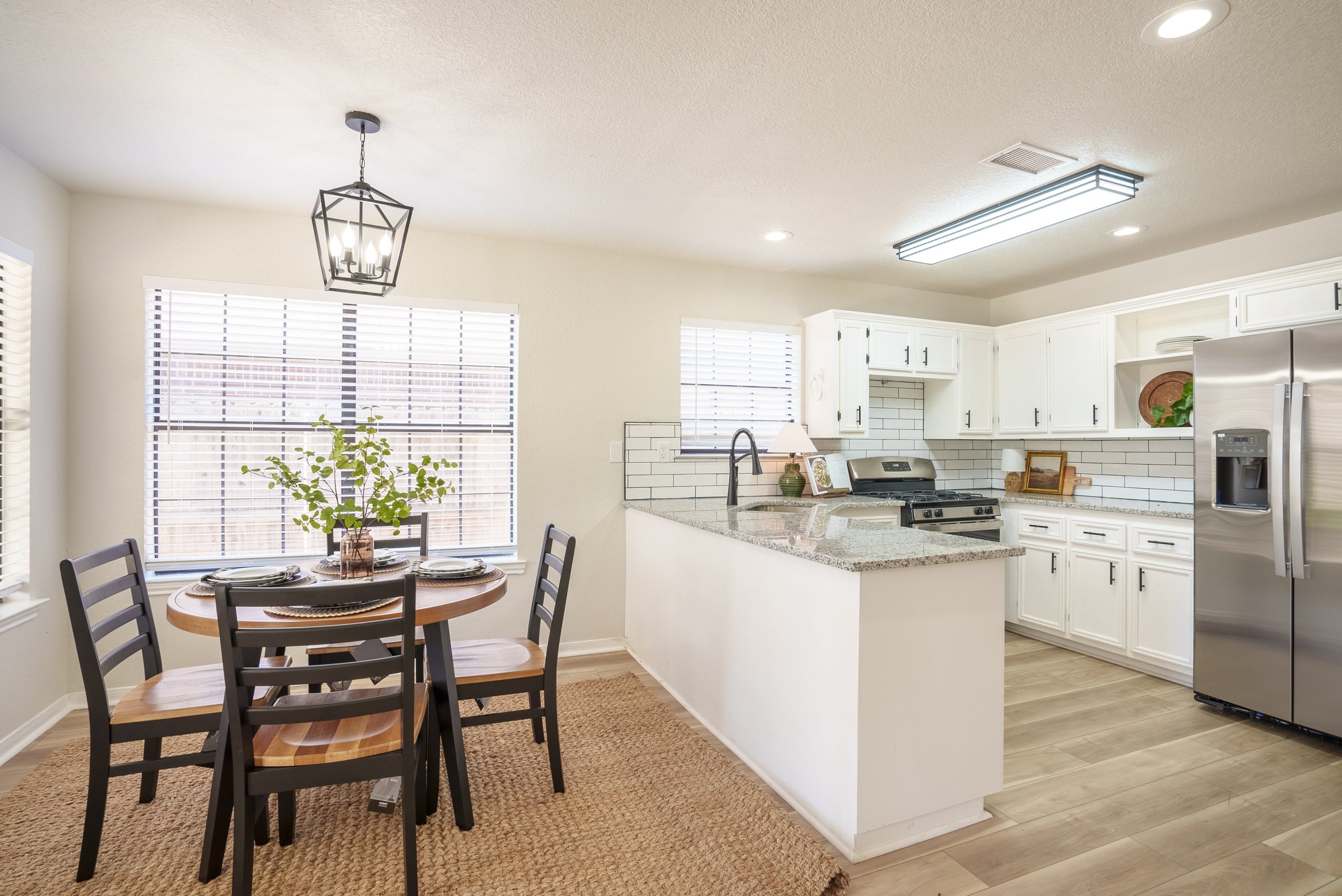 Modern kitchen and dining area with white cabinetry, stainless steel appliances, a granite countertop, and a wooden dining table with four chairs. A decorative pendant light hangs over the table, and house plants add greenery to the space.