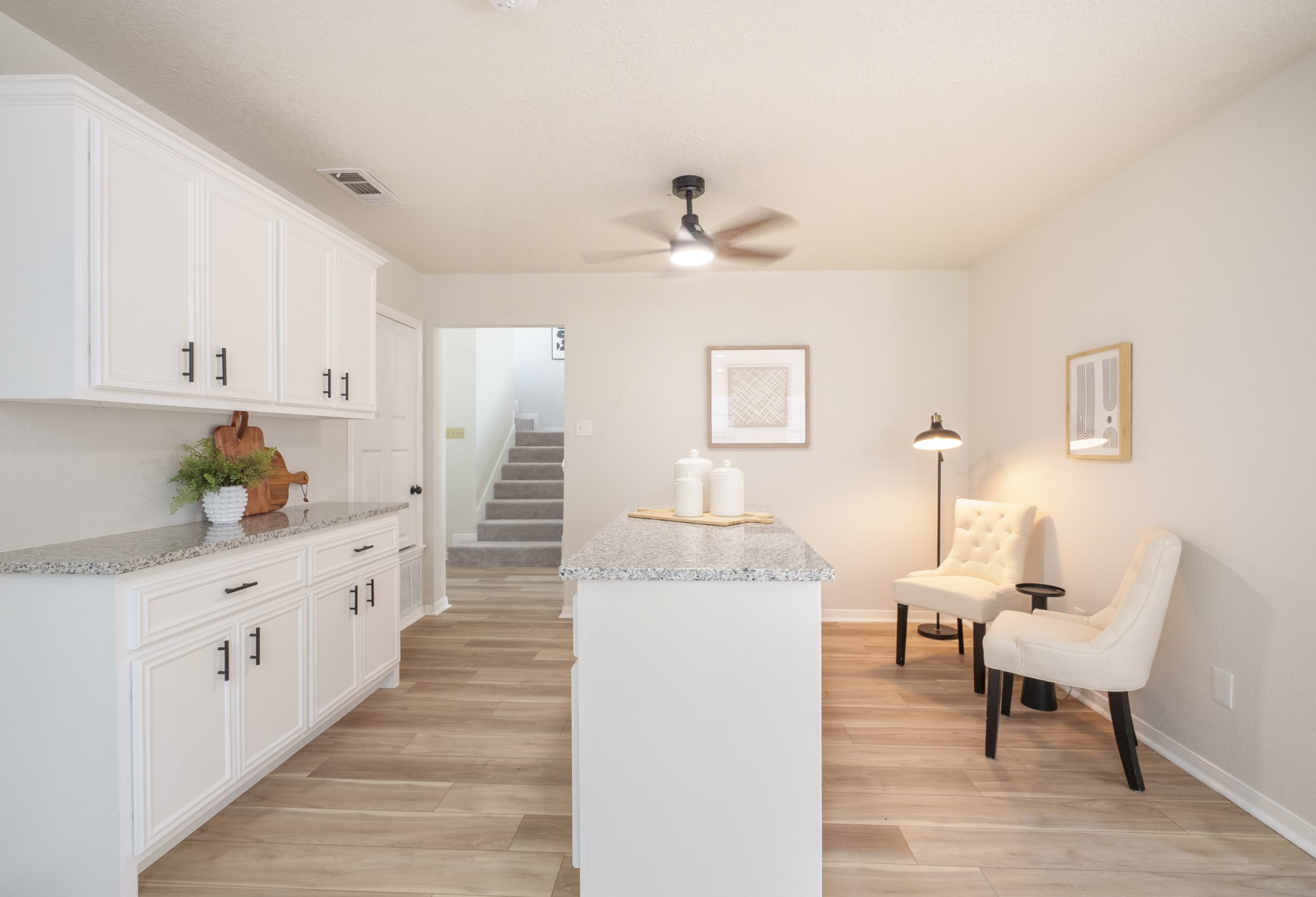 Modern kitchen with white cabinets, granite countertops, wooden flooring, two white upholstered chairs, wall art, and a staircase in the background.