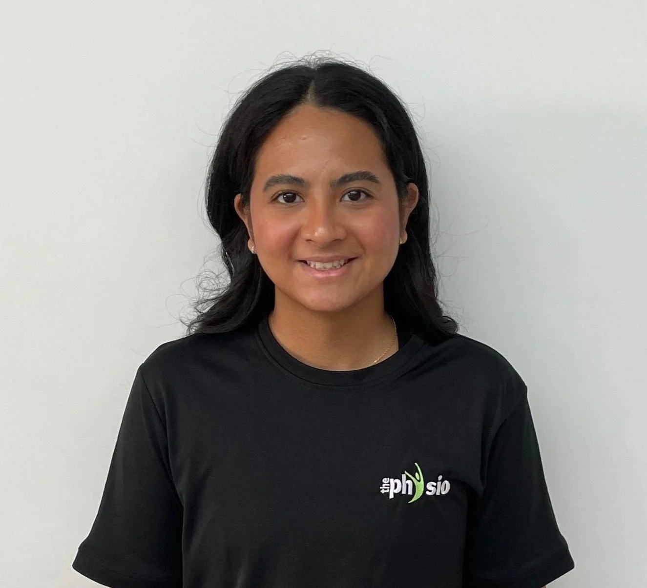 A young woman with dark hair and brown skin smiling, wearing a black t-shirt with a logo reading "the physio" on a plain white background.