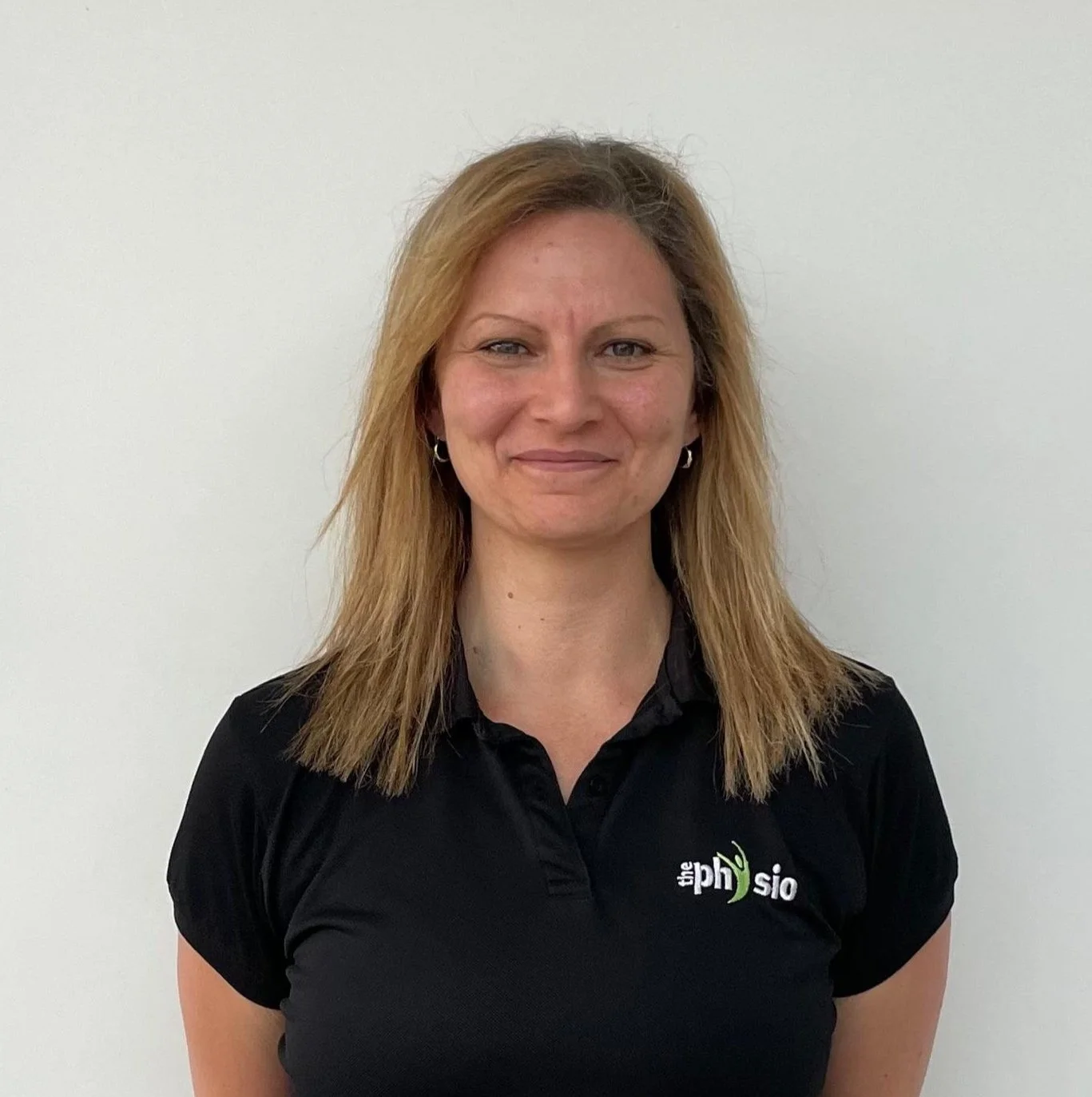 A woman with shoulder-length blonde hair, wearing a black polo shirt with a logo on the chest that says 'the physio', standing against a plain white wall.