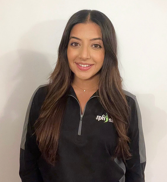 Young woman with long wavy brown hair smiling, wearing a black quarter-zip athletic shirt with gray accents and a small necklace, standing against a plain white wall.