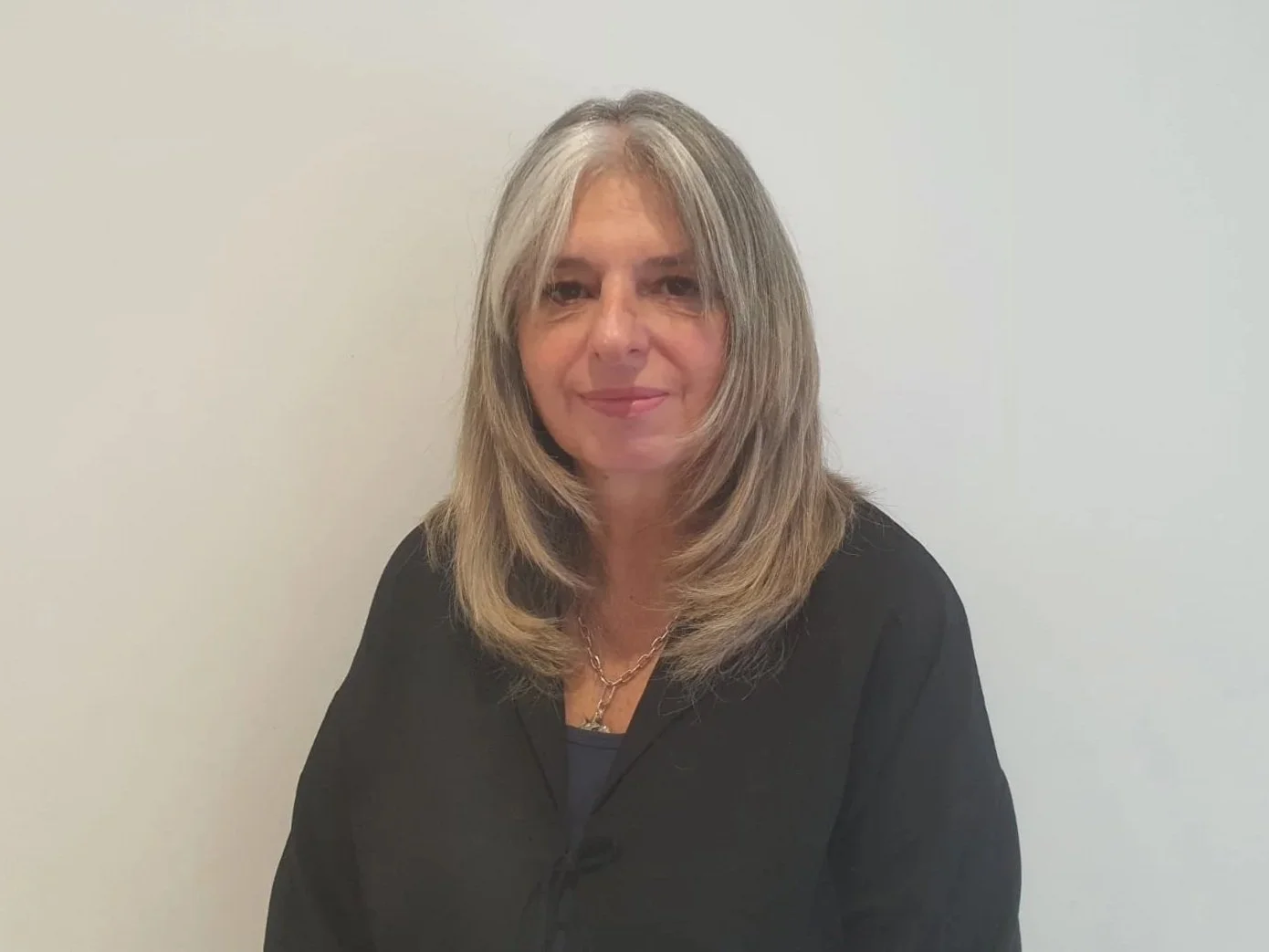 A woman with shoulder-length gray hair, wearing a black blazer and a necklace, standing against a plain white wall.