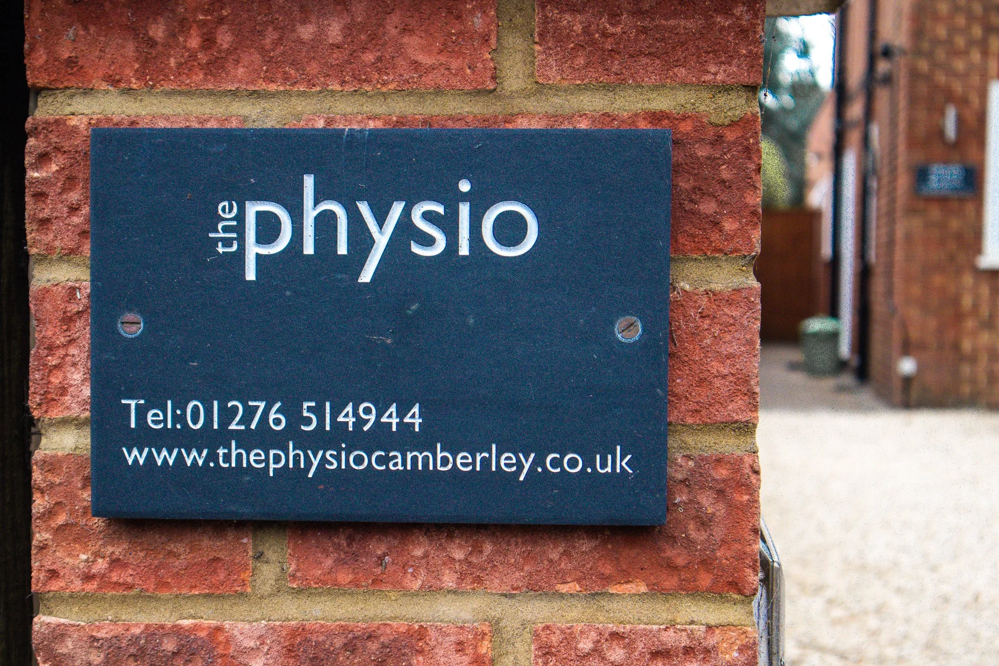 A dark blue sign on a brick wall that reads 'the physio', with contact information including a phone number and website.