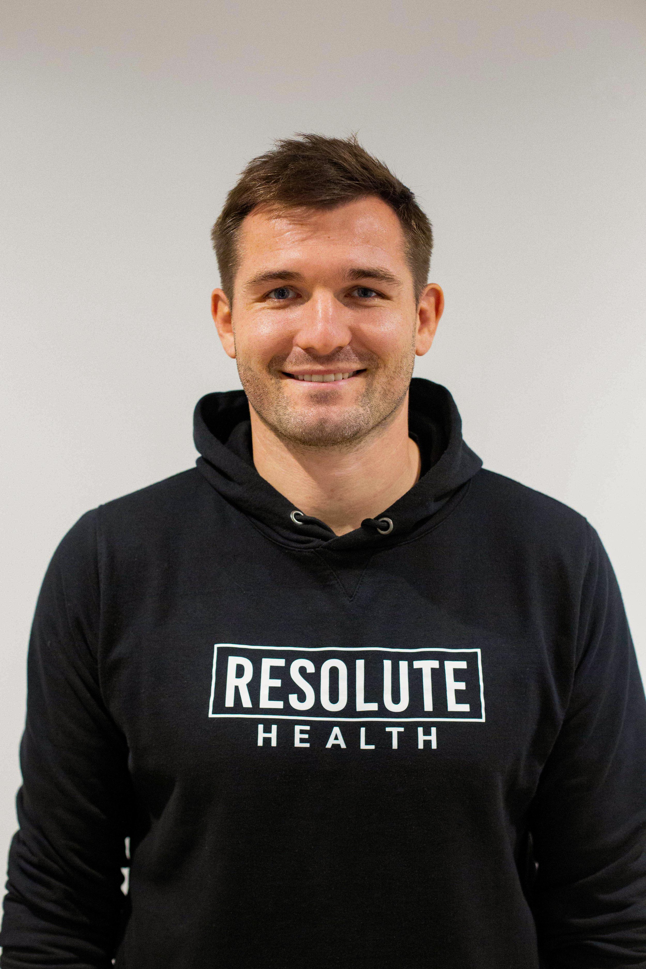 A young man smiling, wearing a black hoodie with 'RESOLUTE HEALTH' printed on it, standing against a plain white background.