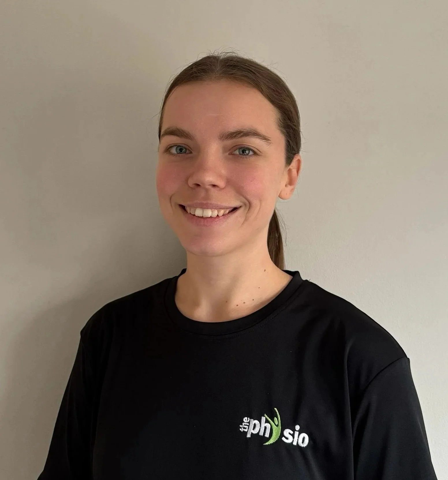 Portrait of a young woman with brown hair, smiling, wearing a black shirt with the logo 'the physi' visible on the chest, standing against a plain light-colored wall.