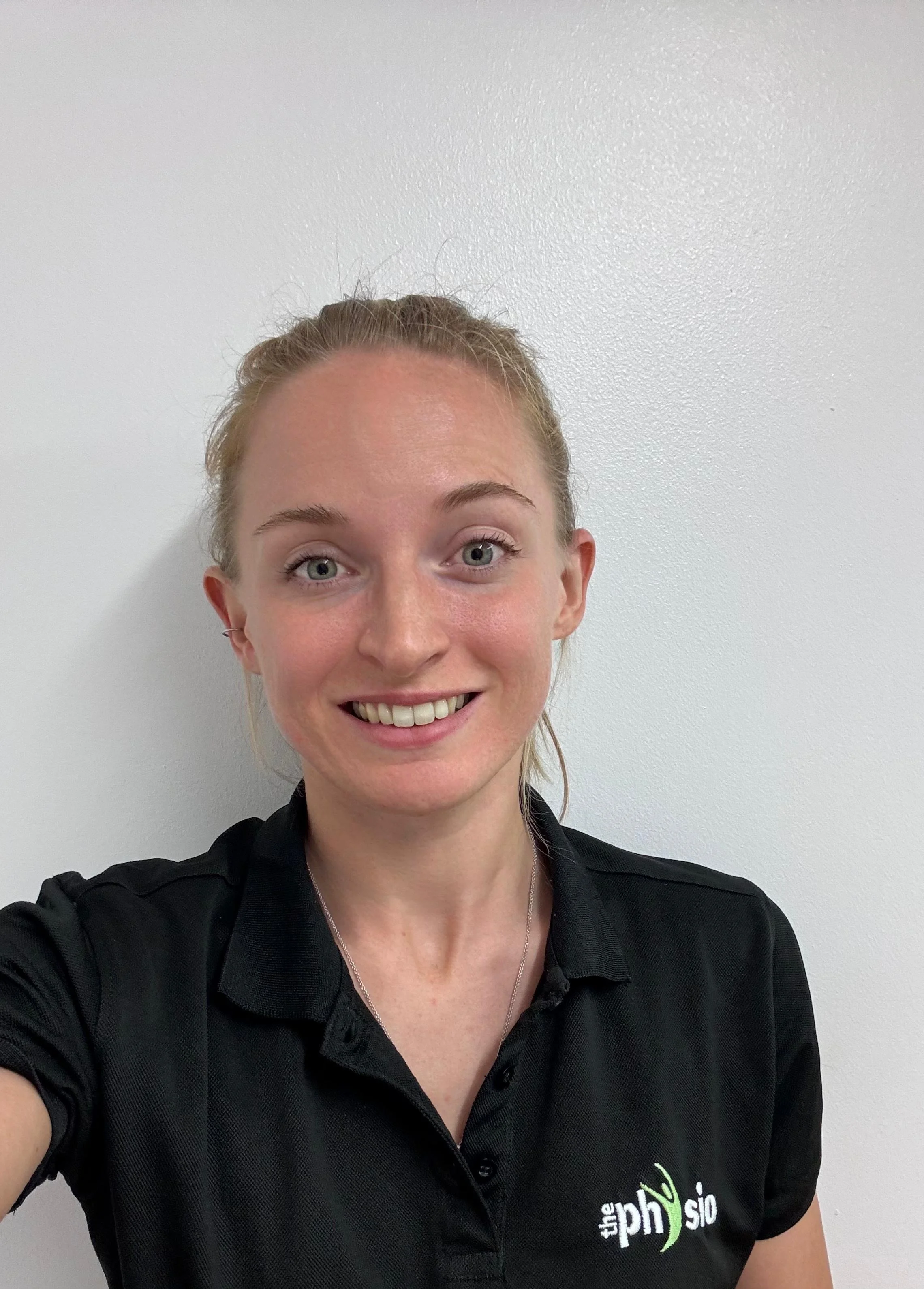 A young woman with blonde hair tied back, smiling, wearing a black polo shirt with a logo on the left side, standing against a white wall.