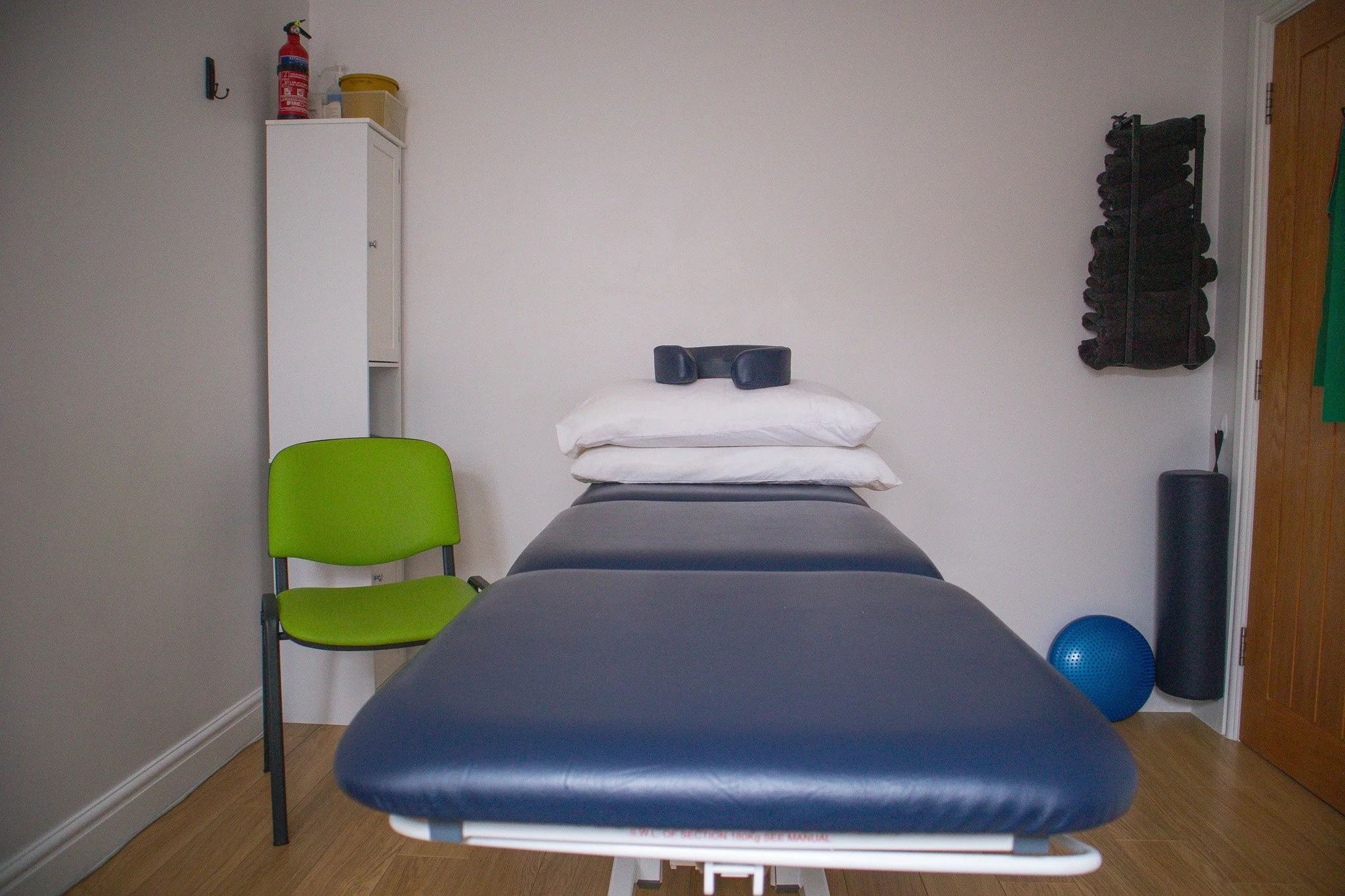 Massage table with stacked pillows, a green chair on the left, and fitness equipment including a blue exercise ball and black foam roller on the right in a therapy room.