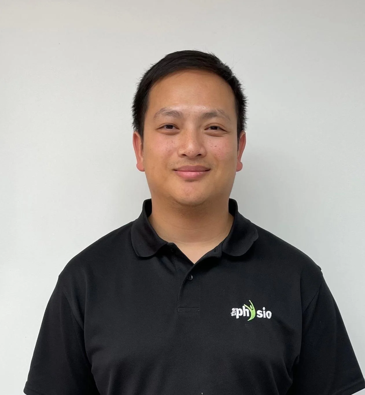 A man wearing a black polo shirt with a logo that says 'the physio' standing against a plain wall.