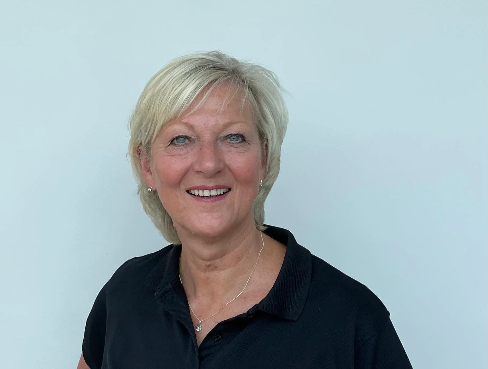 An older woman with short blonde hair smiling, wearing a black polo shirt and a necklace with a pendant, standing against a plain light-colored background.