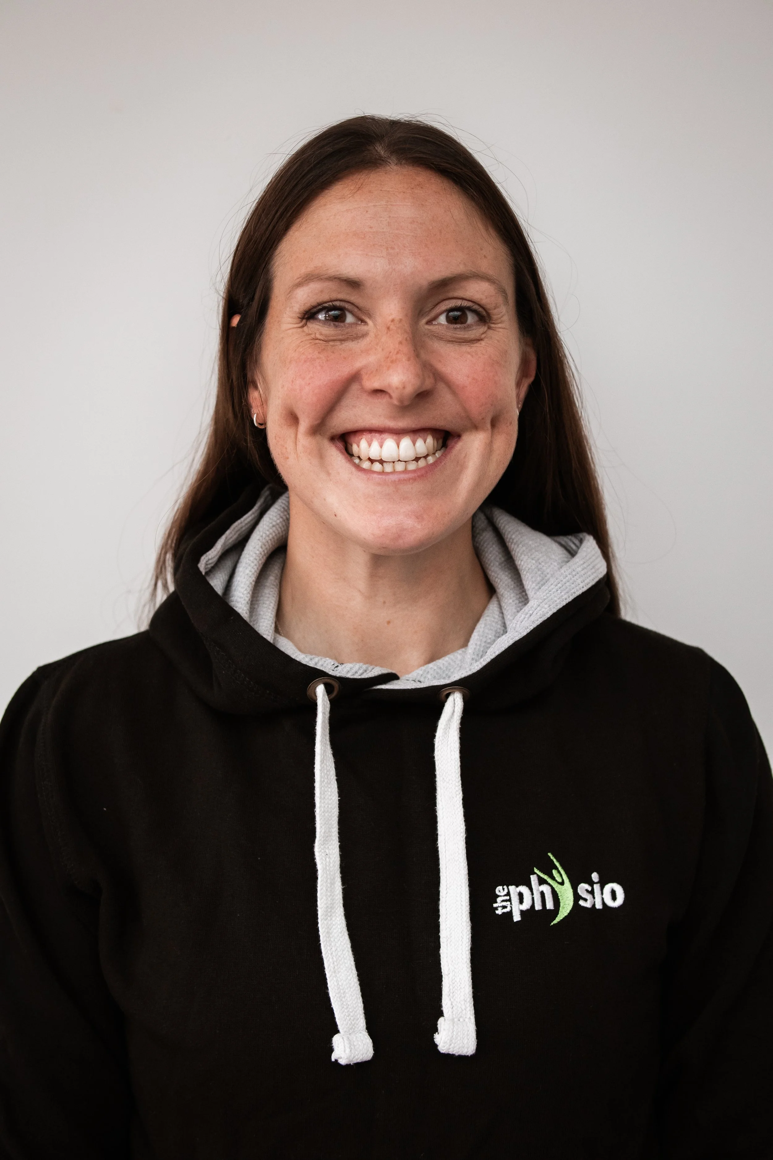 A woman smiling, wearing a black hoodie with a logo that says 'the physio' in white and green text, with a gray hoodie underneath, standing against a plain white wall.