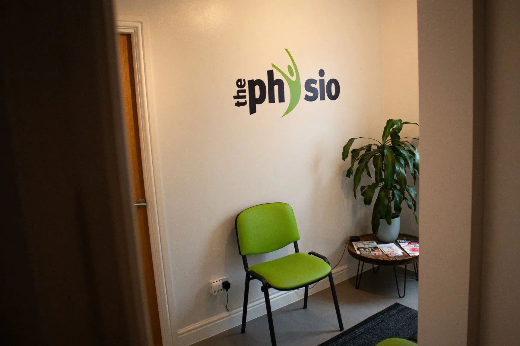 Reception area with a green chair, a potted plant, and a small table with magazines. Wall decal reads 'the physio'.