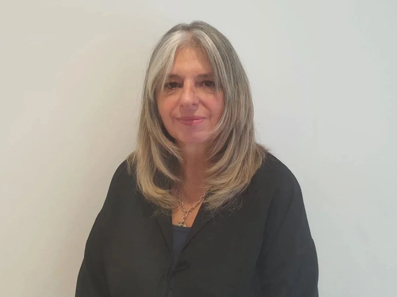 A middle-aged woman with shoulder-length blonde hair posing against a plain white wall, wearing a black top and a gold necklace.