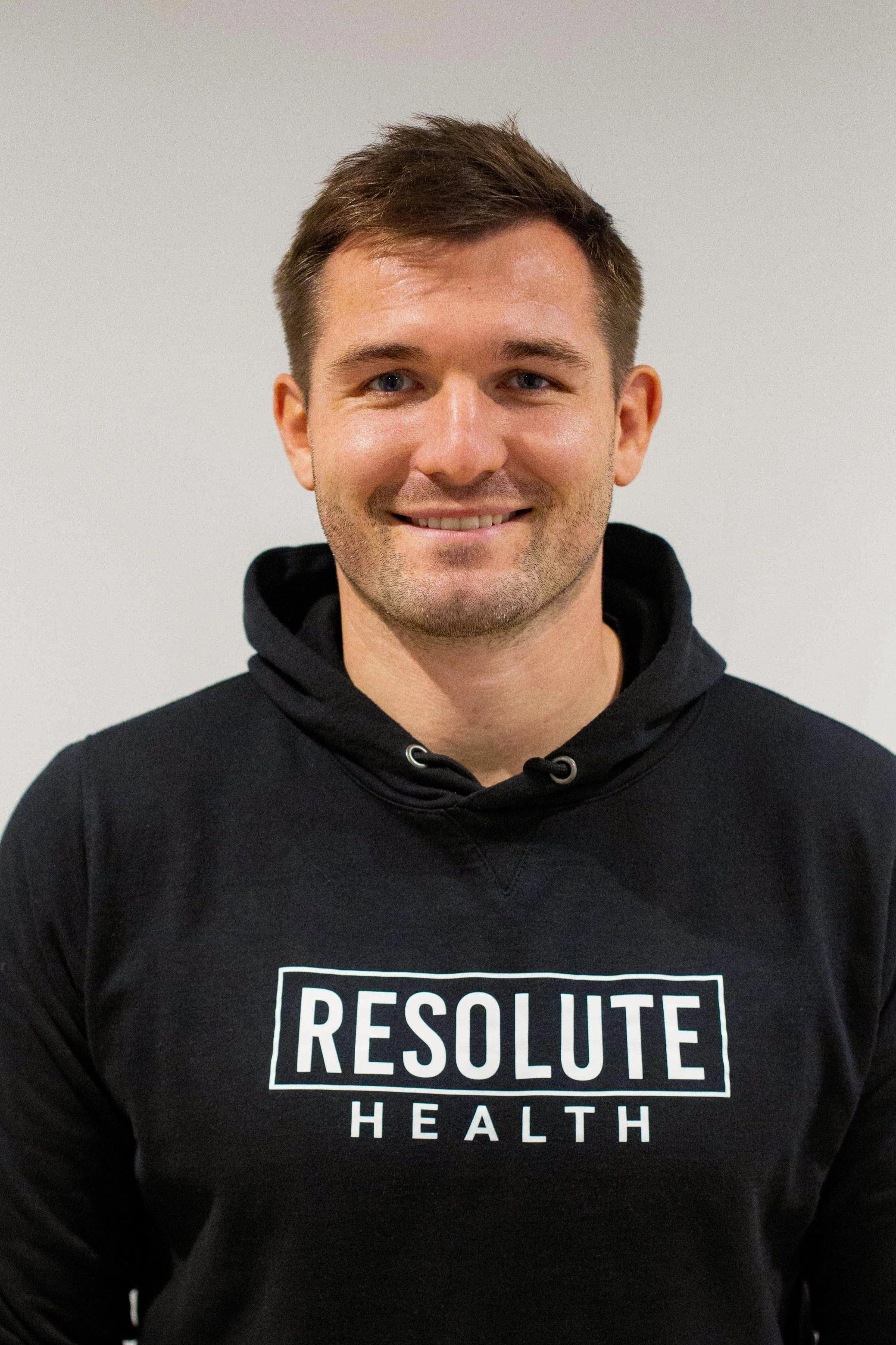 A smiling man wearing a black hoodie with the words 'Resolute Health' printed on it, standing against a plain light-colored background.