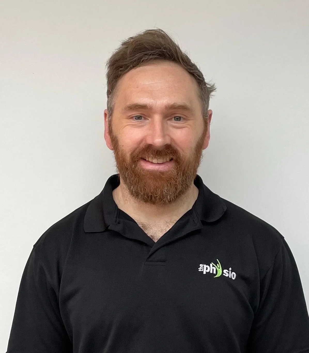 A man with a beard and short brown hair, smiling, wearing a black polo shirt with a logo on the chest that reads 'the physio' with a green checkmark.