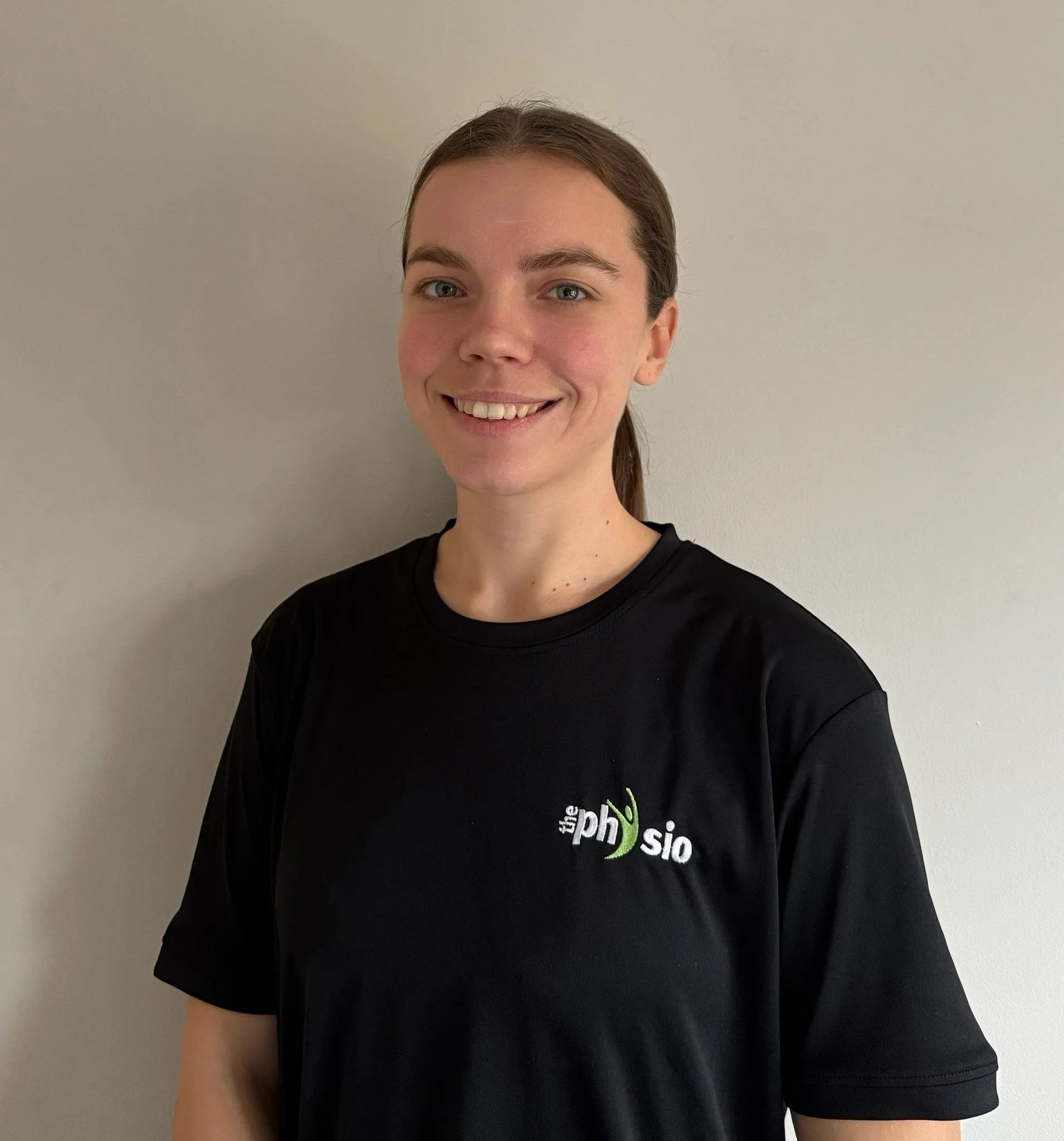 A young woman with brown hair tied back, smiling, wearing a black T-shirt with a logo on the left side of her chest that reads 'the physio' with a green and white design, standing against a plain light-colored wall.