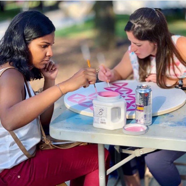 Two women painting geometric designs on a circular canvas at an outdoor table, with art supplies and drinks nearby.