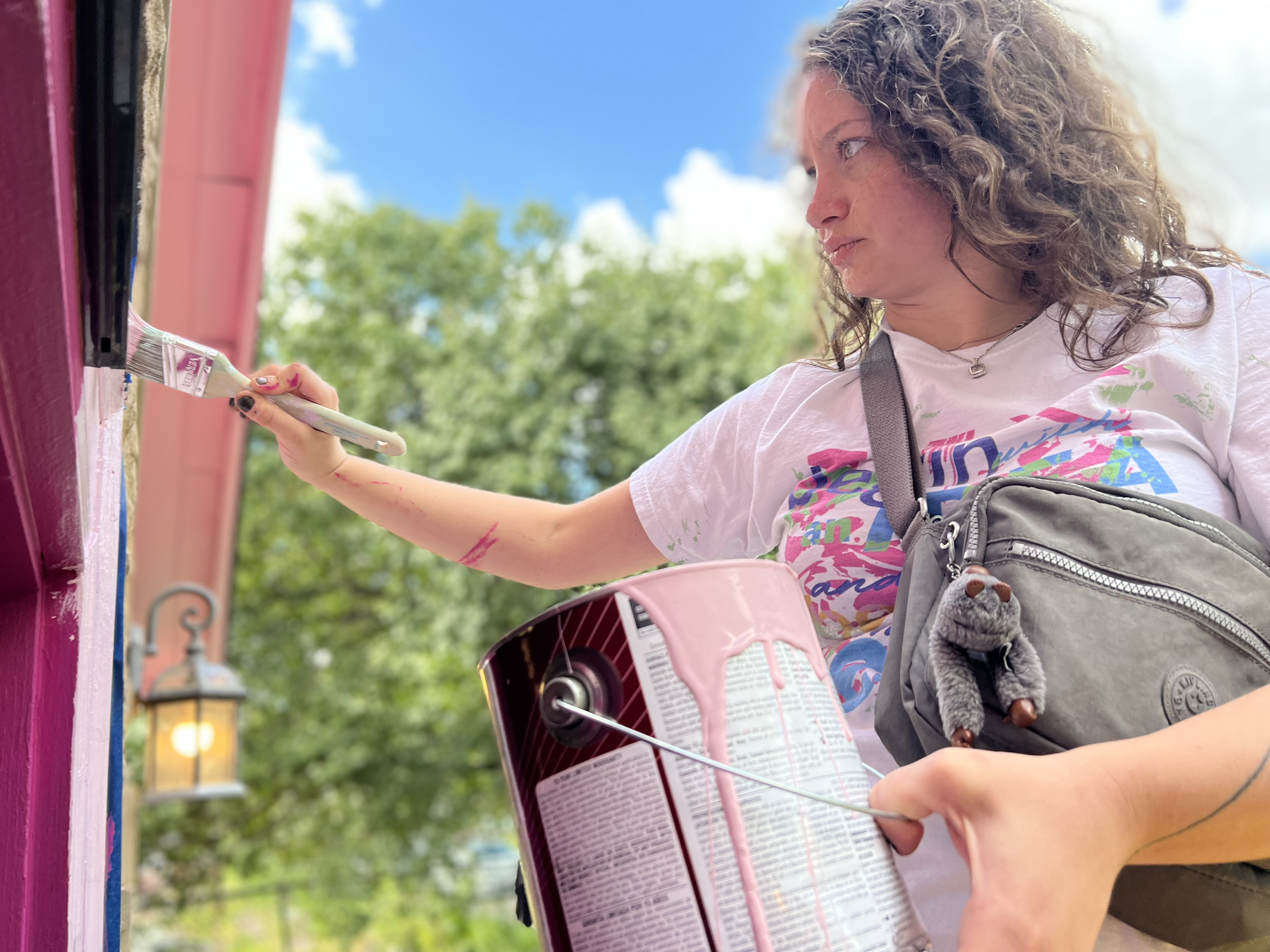Person painting a wall with pink paint, holding a paint can and brush, wearing a white t-shirt and gray bag, outdoors.