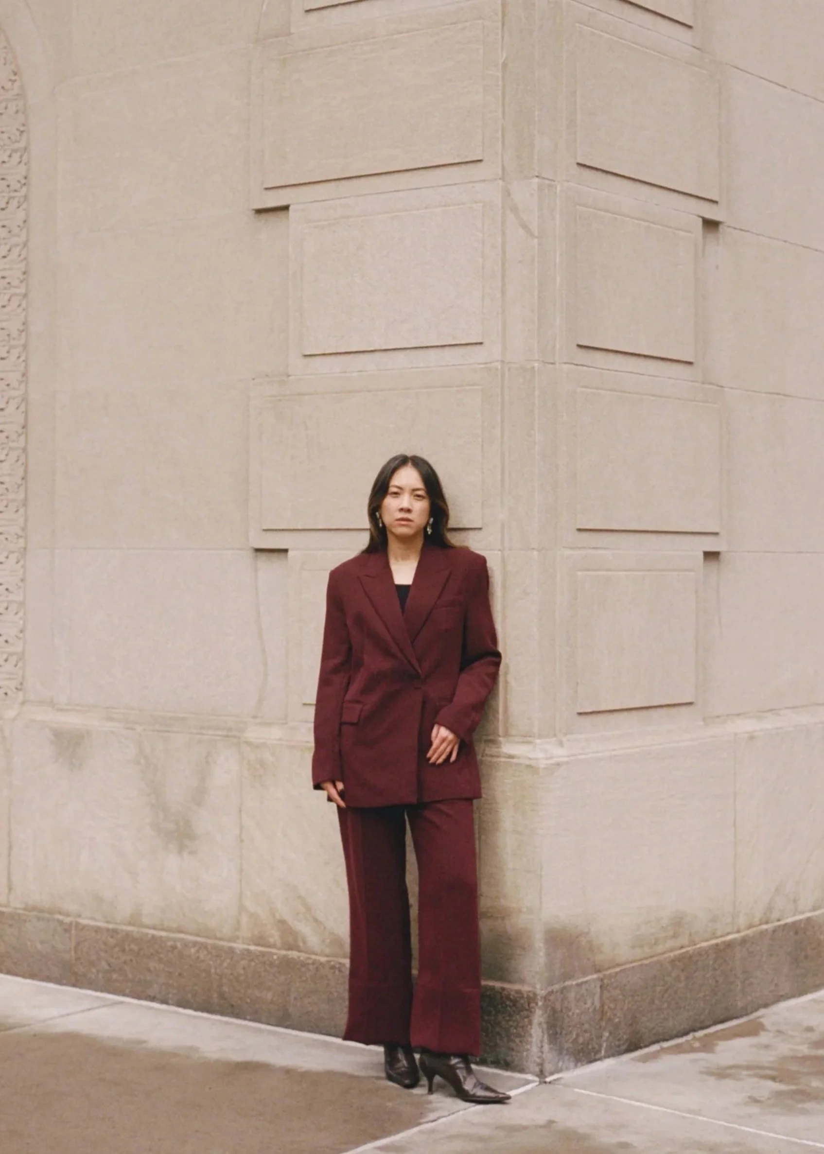 A woman standing in front of a beige stone building, wearing a dark red suit with a blazer and wide-leg pants, and black heeled boots.