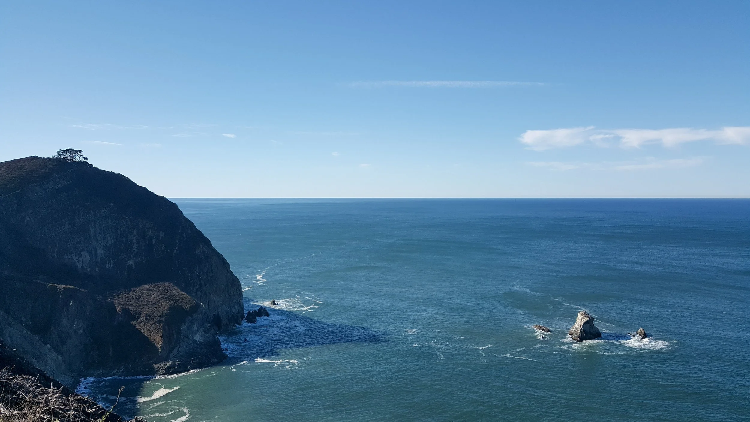 A coastal landscape showing a cliffside with trees on top, rocky formations in the ocean, and a clear blue sky with a few clouds.