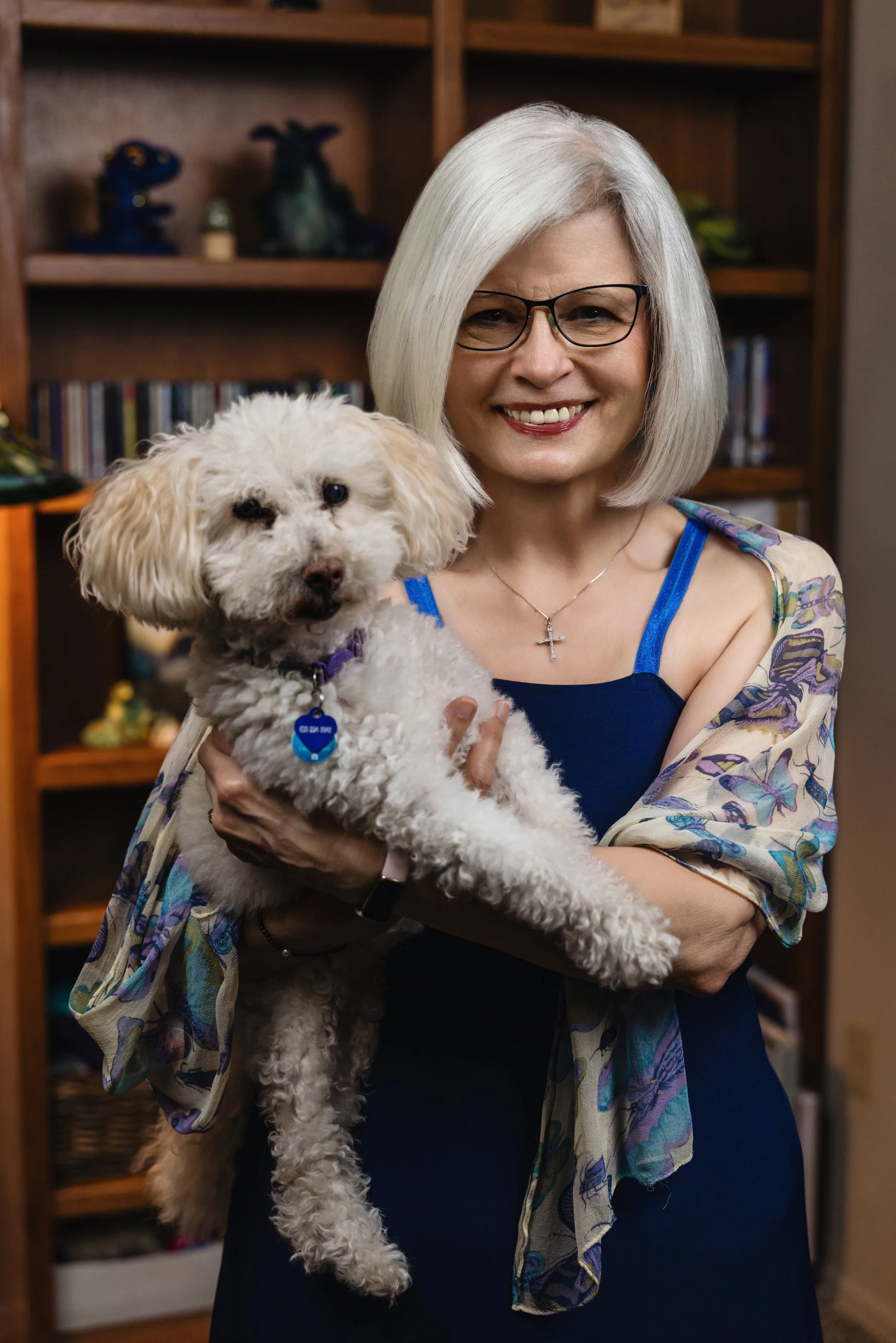 KT Sweet in a blue dress holding a small, curly-haired white dog in a room with a wooden bookshelf in the background.