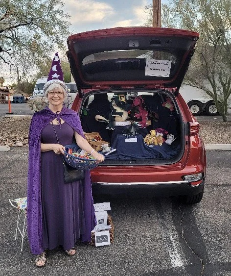 Woman dressed as a witch, wearing a purple dress, cape, and a pointed hat with stars, standing next to an open vehicle trunk filled with stuffed animals, decorations, and bags in a parking lot.