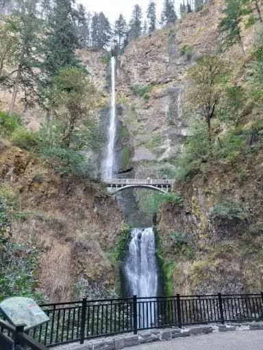 A scenic view of a waterfall cascading down a rocky cliff, with a bridge crossing in front of it and a viewpoint railing at the bottom.