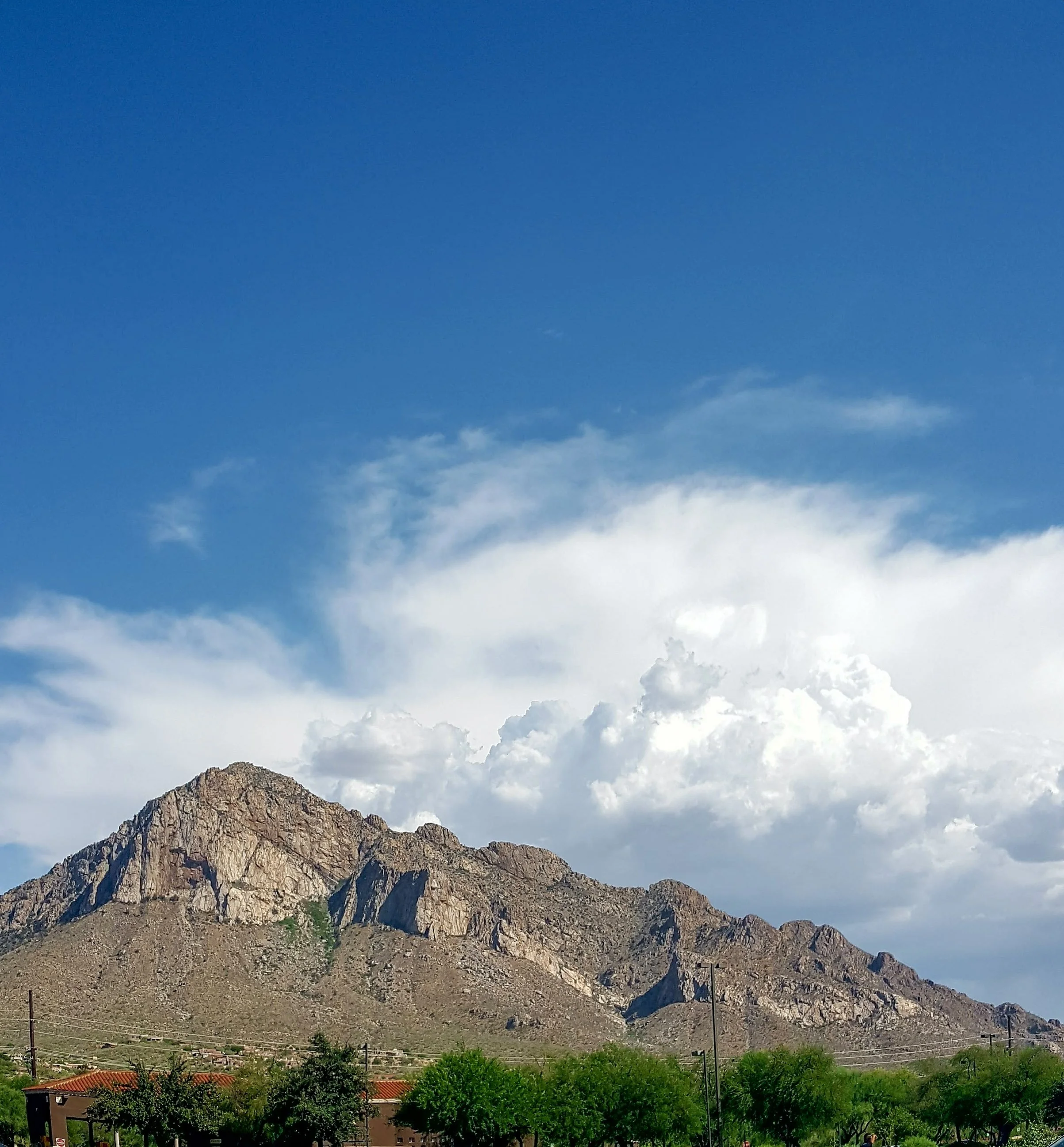 A mountain range under a partly cloudy sky with green trees and some buildings at the base.