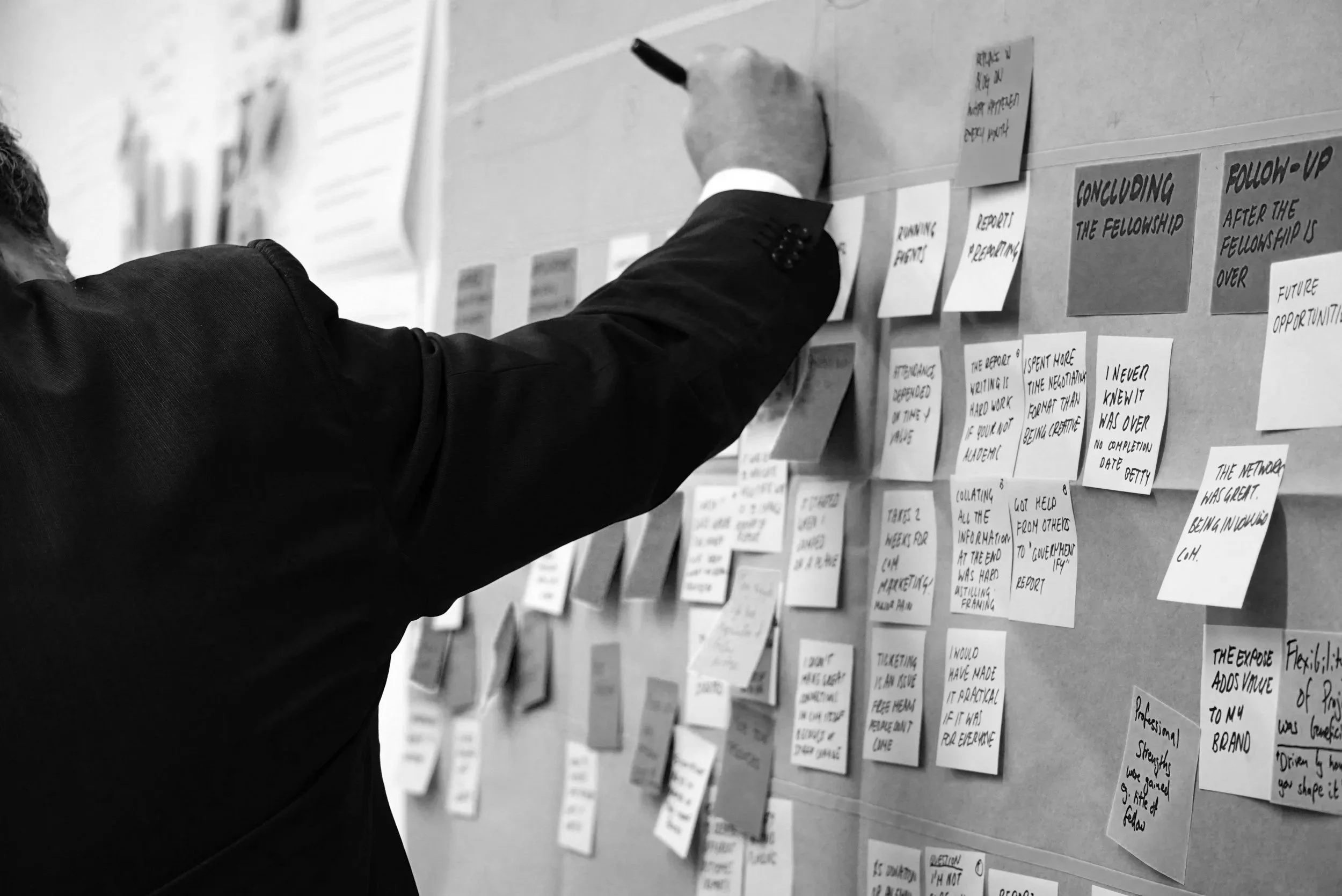 A person writing on a sticky note on a wall covered with many other sticky notes containing handwritten notes.