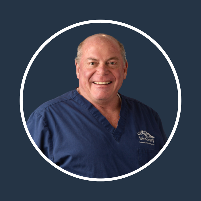 Smiling person in navy blue scrub top with "McKinley" text on left chest, set against a dark background with white circular border.