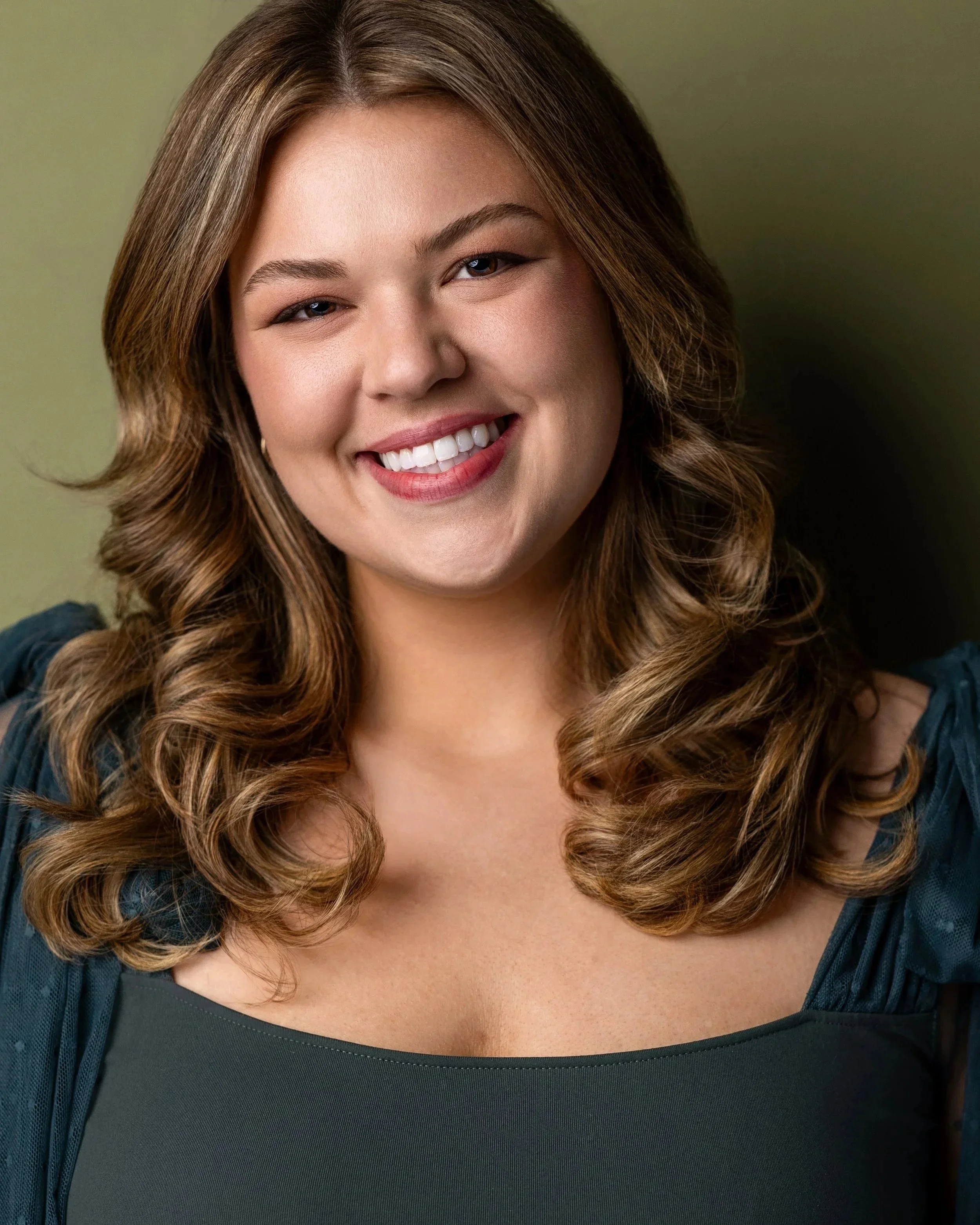 Kat's headshot shows her with shoulder-length, wavy brown hair wearing a dark green top against a muted green background.