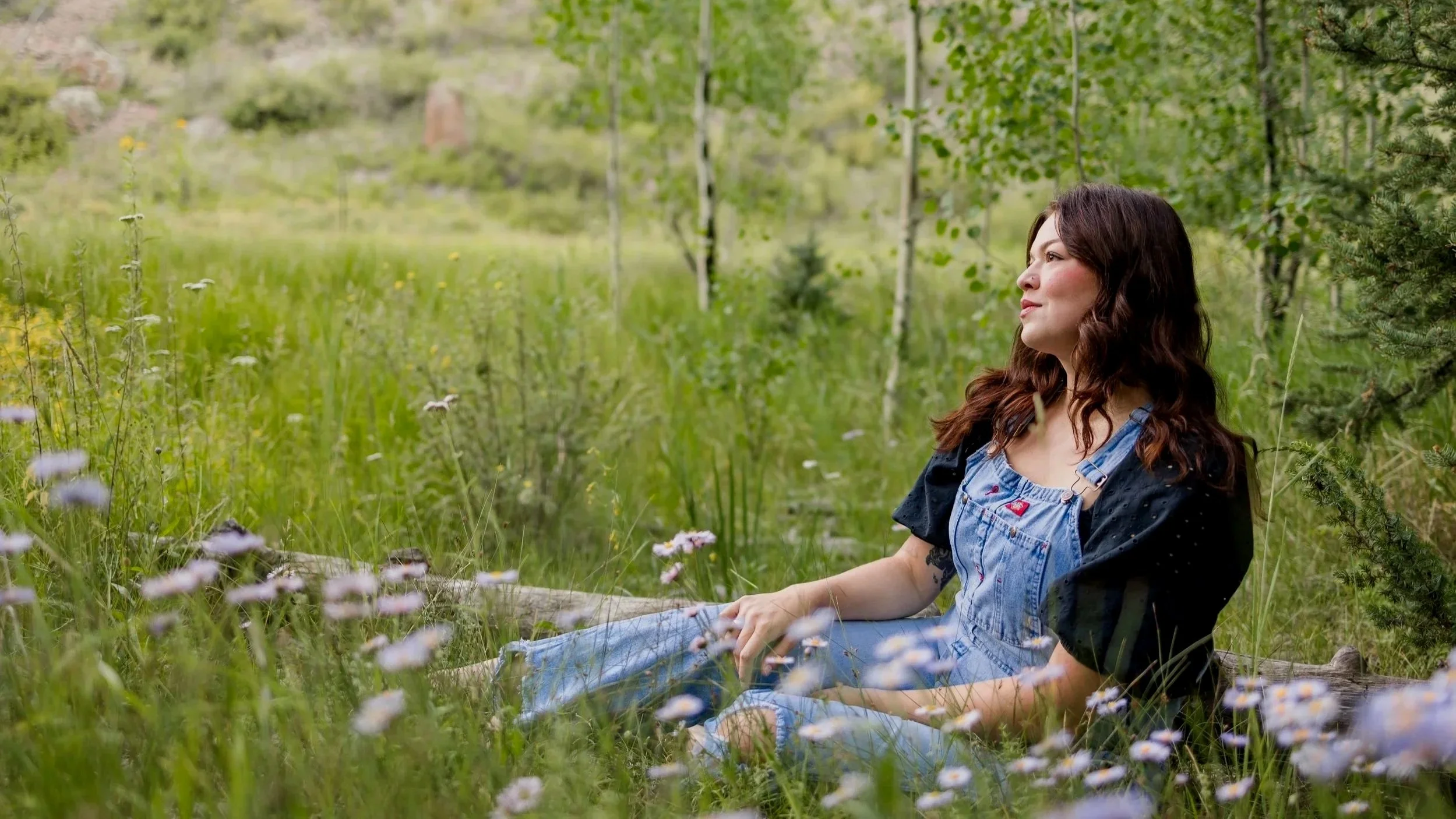 Kat, a woman with long brown hair sitting in a grassy meadow surrounded by wildflowers, wearing denim overalls and a black short-sleeve shirt.