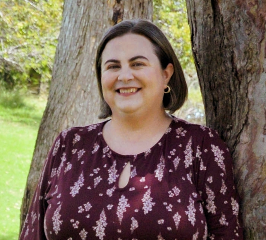 A woman smiling outdoors, standing next to a large tree, wearing a patterned burgundy blouse with a keyhole neckline. Green foliage and grass are in the background.